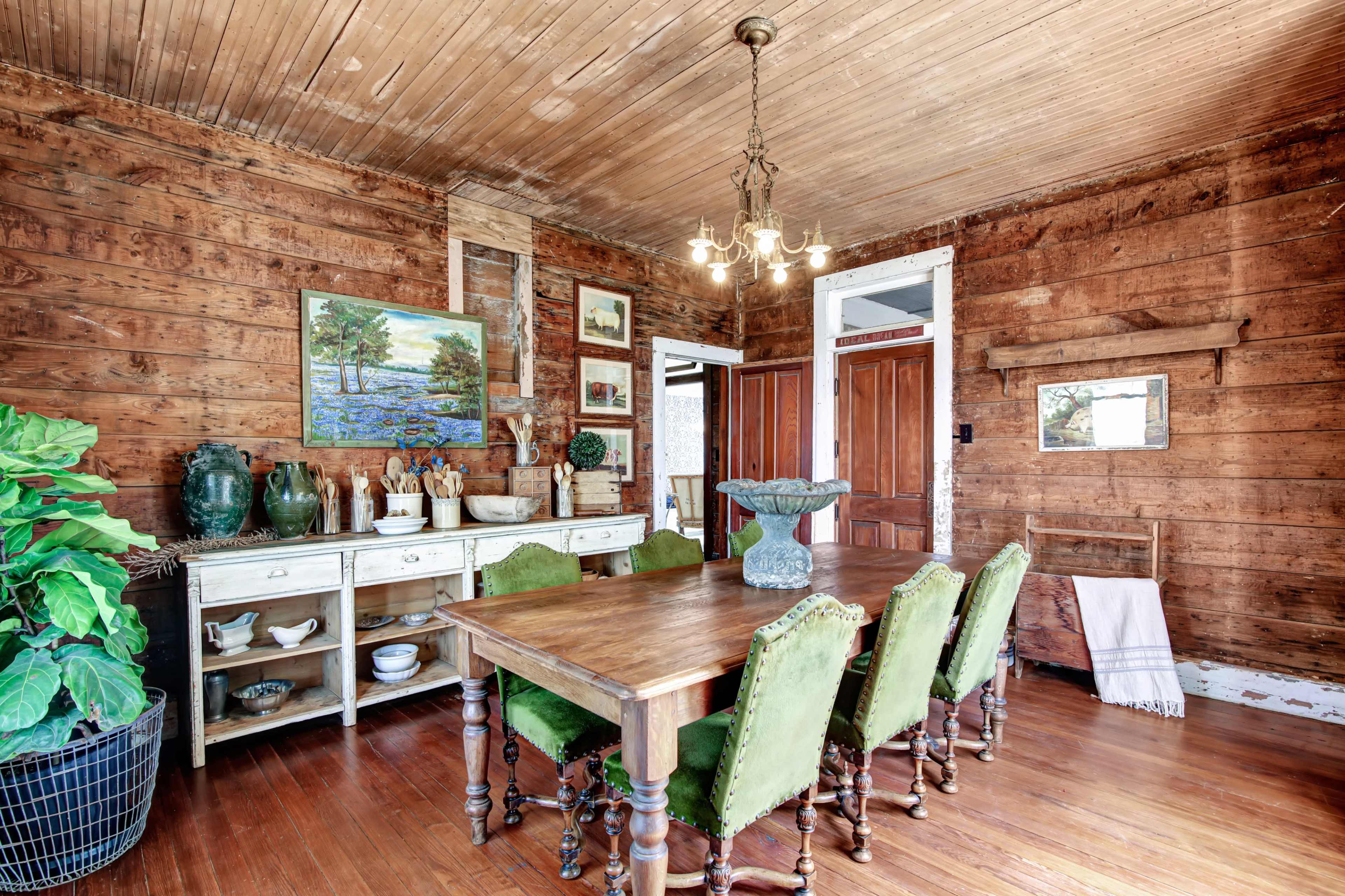 A wooden dining room features a large table surrounded by green chairs, with a sideboard displaying various items against a backdrop of rustic wooden walls.