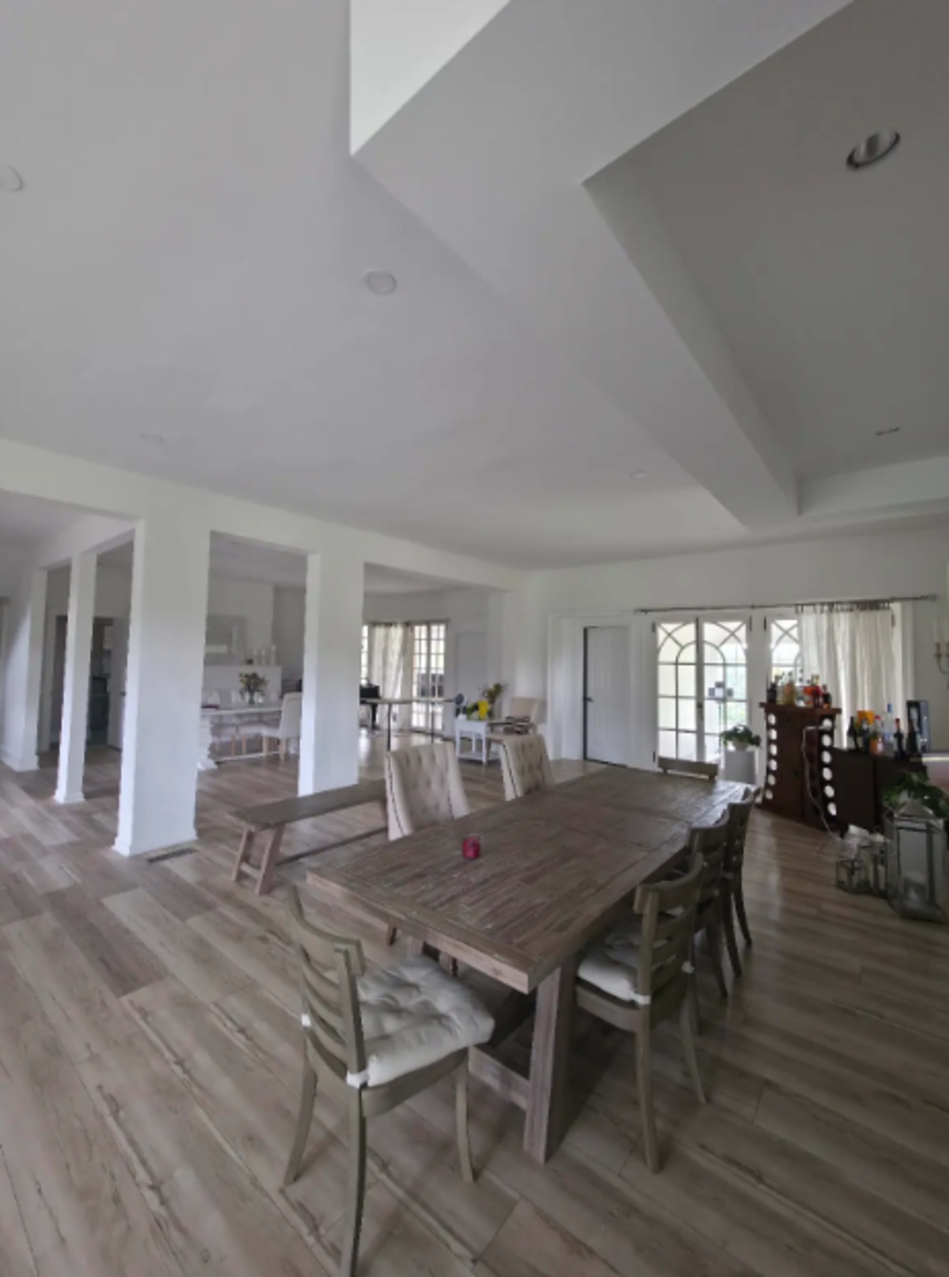 The image shows a spacious, modern dining area featuring a large wooden table surrounded by chairs, with large windows and light-colored walls.