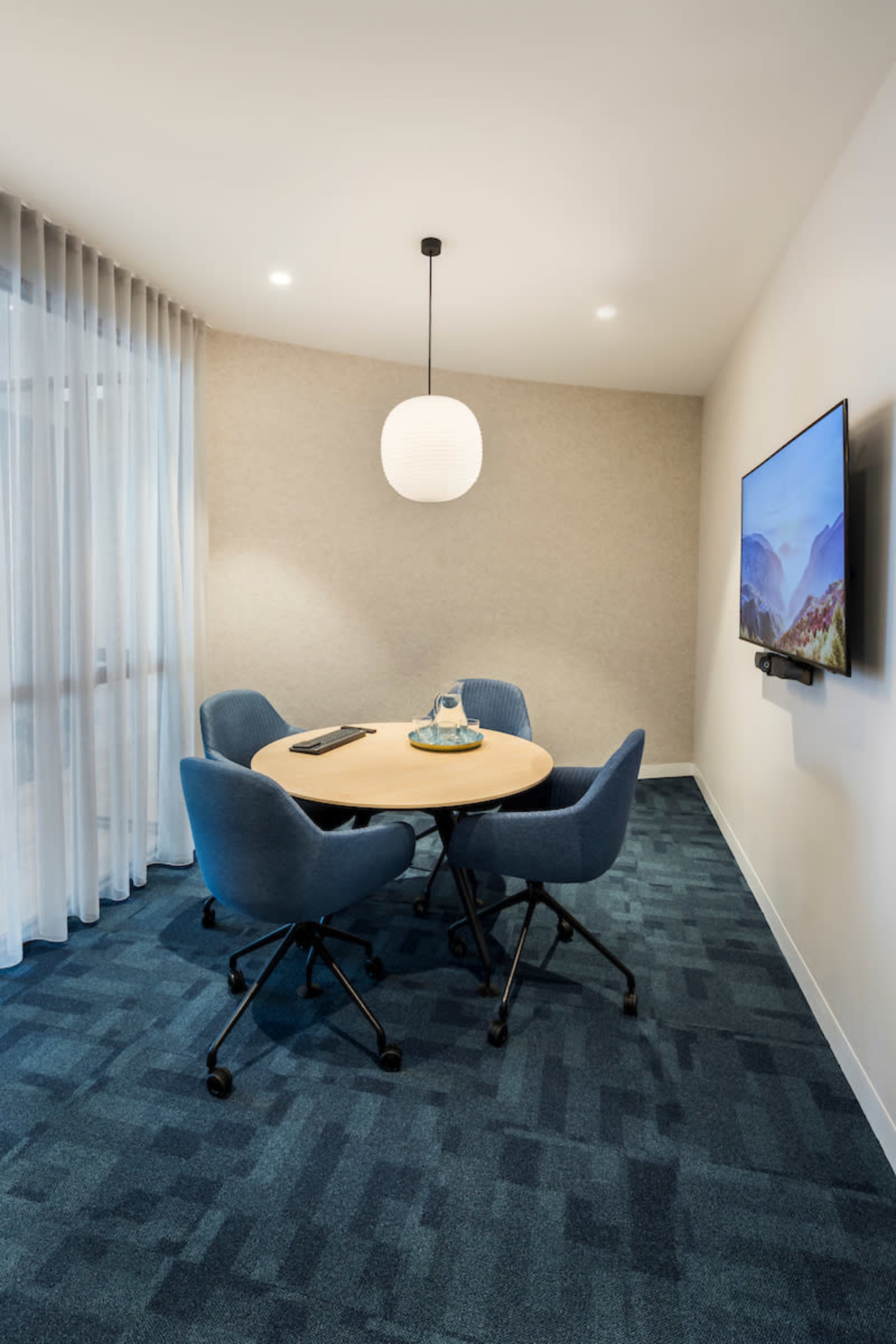 A modern meeting room features a round wooden table surrounded by four blue chairs and a wall-mounted television.