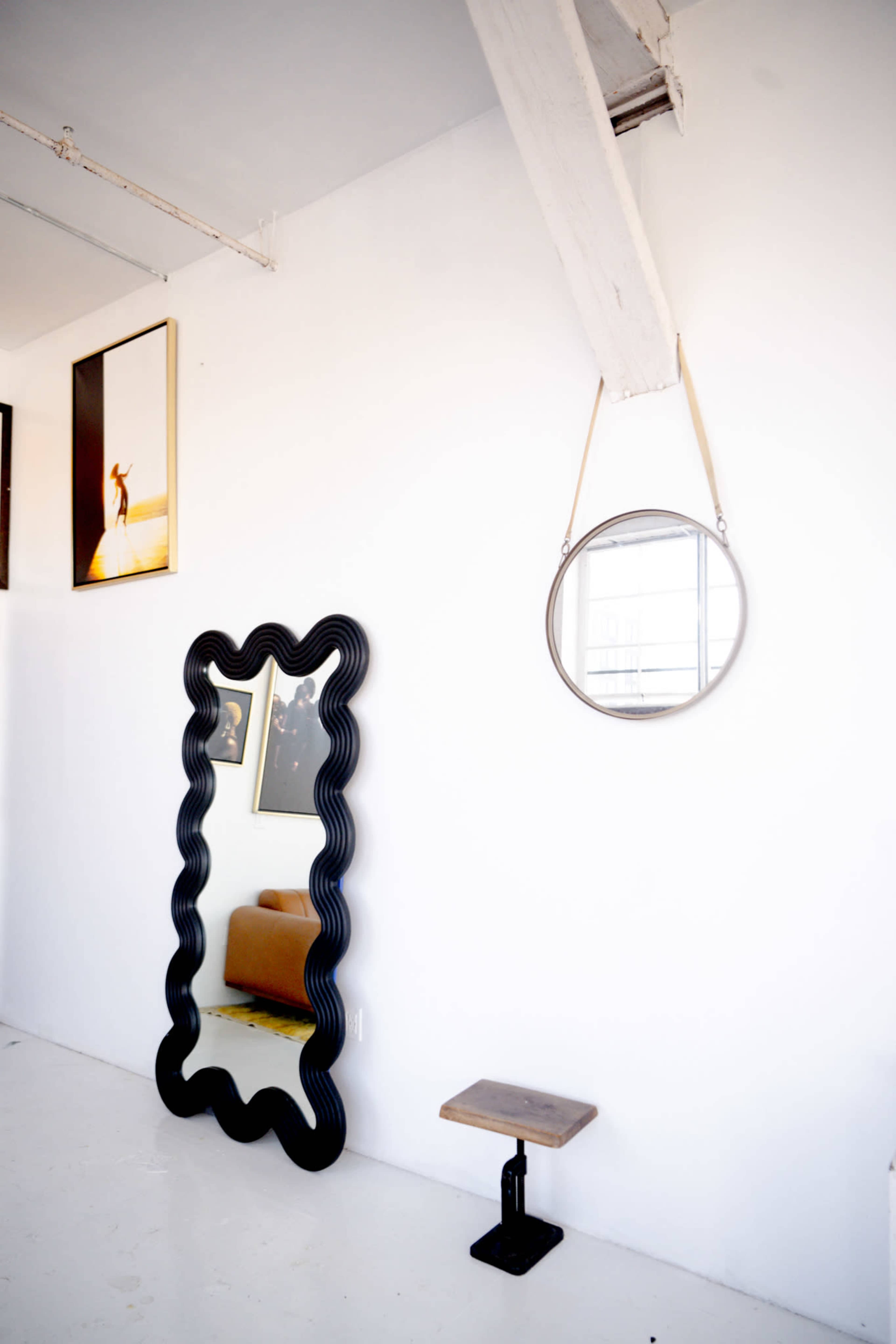 A black wavy-framed mirror, a round mirror hanging from a strap, a small wooden stool, and two framed artworks on a white wall.