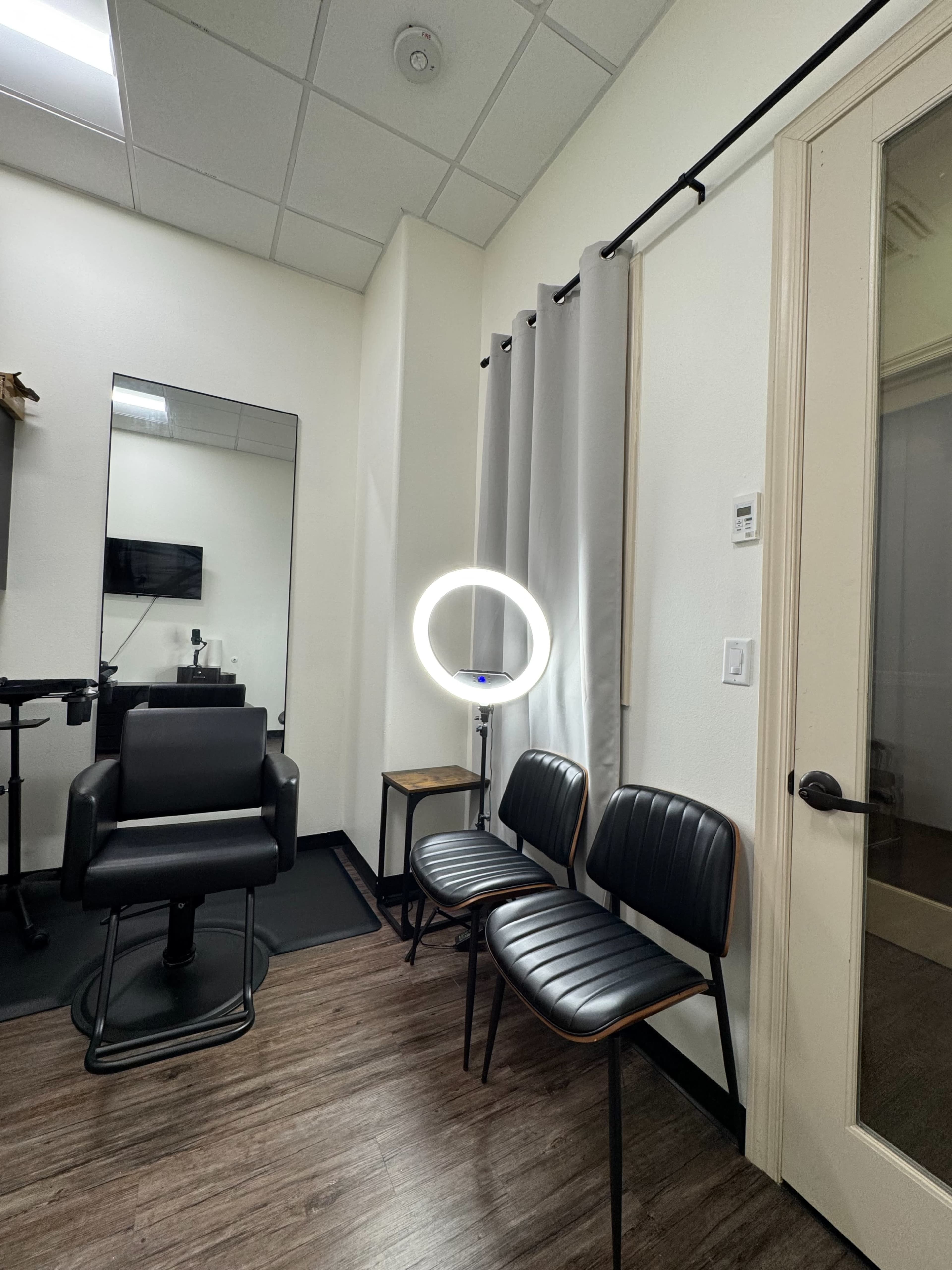 The image shows a small salon or grooming room with a black chair, a mirror, two black chairs, a ring light, and a partially open door.