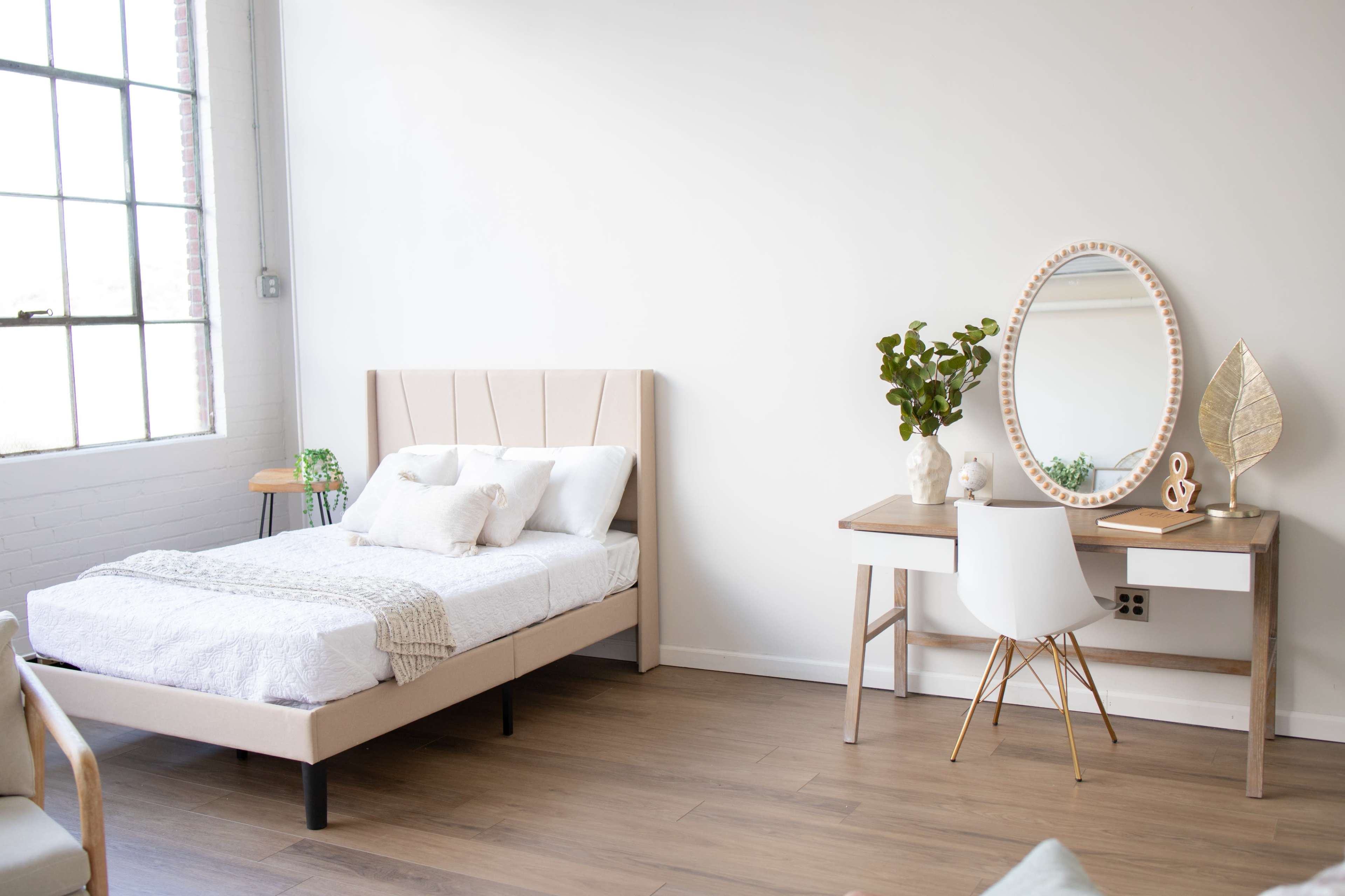A minimalist bedroom featuring a bed with a light-colored headboard, a small wooden nightstand, a desk with a round mirror, and a potted plant.