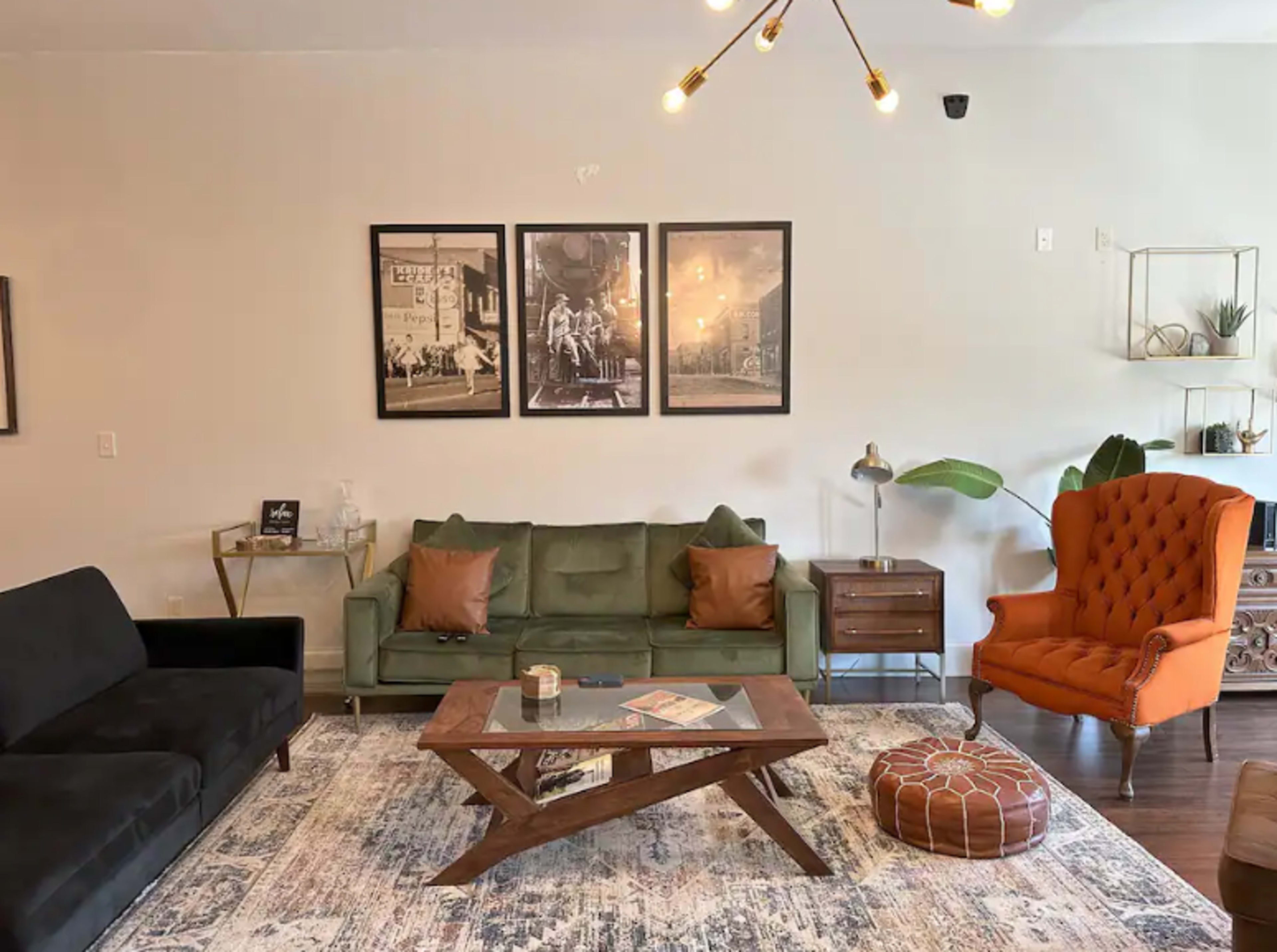 The image shows a living room with a green sofa, a black sofa, an orange armchair, a wooden coffee table, and framed art on the wall.