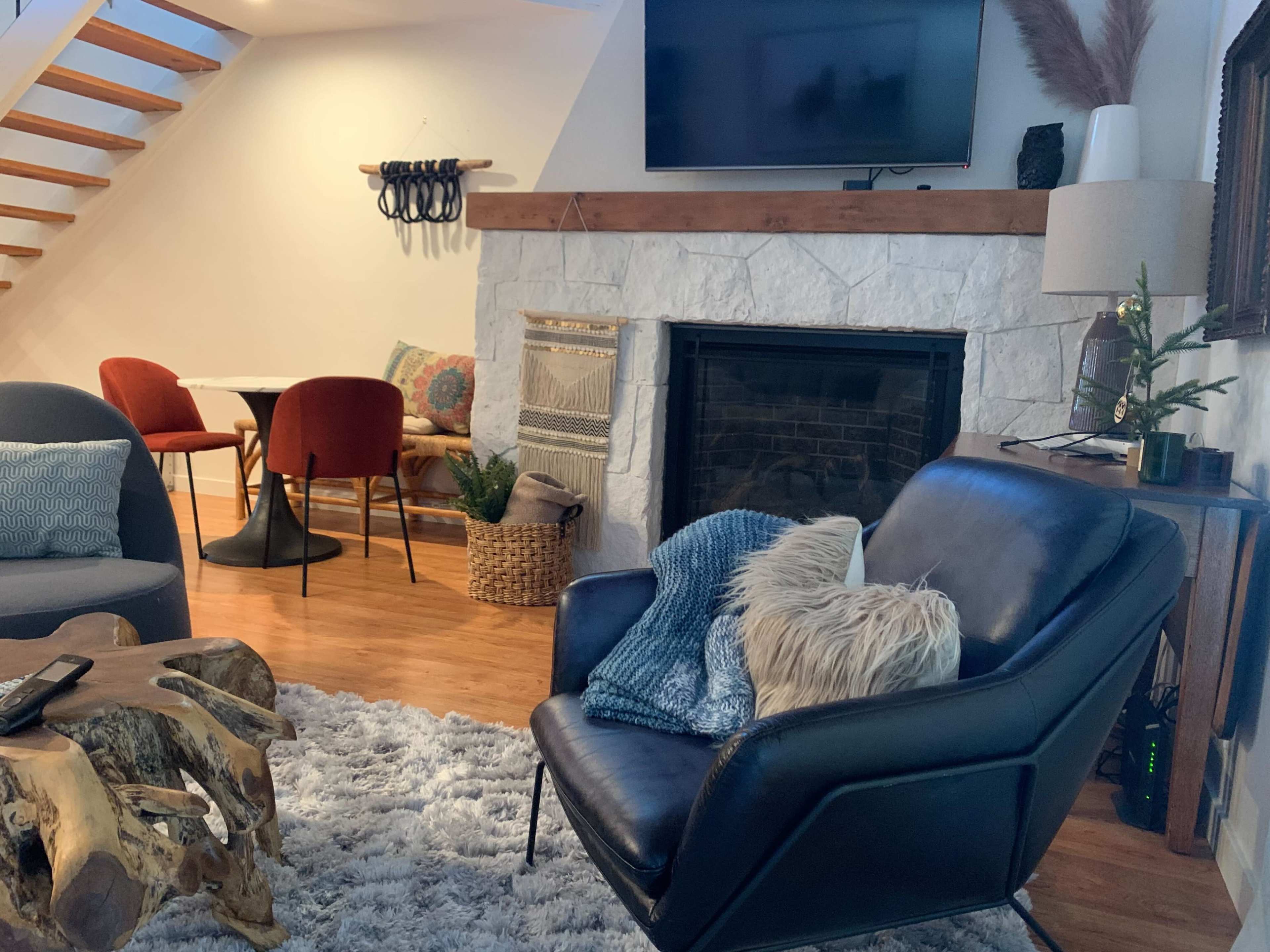 A cozy living space featuring a black leather chair with a throw blanket, a stone fireplace, a wooden table set for two, and a staircase in the background.