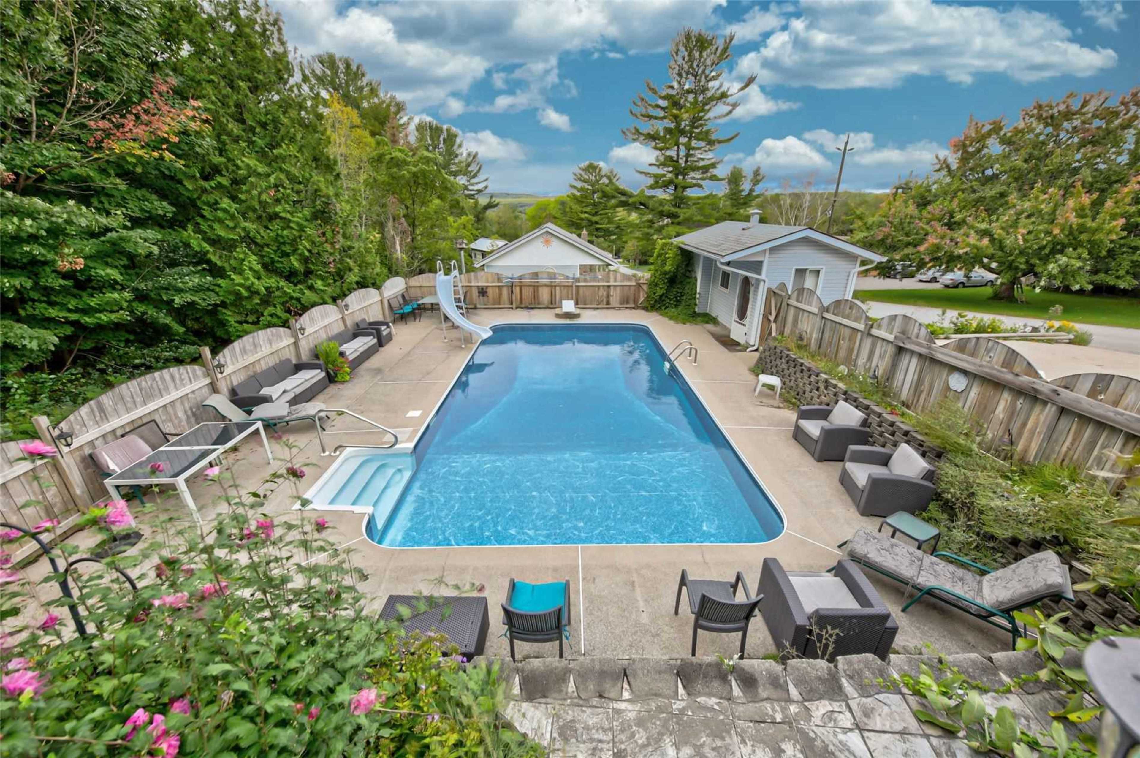 A rectangular swimming pool surrounded by lounge chairs and a slide, with a backdrop of trees and a house.