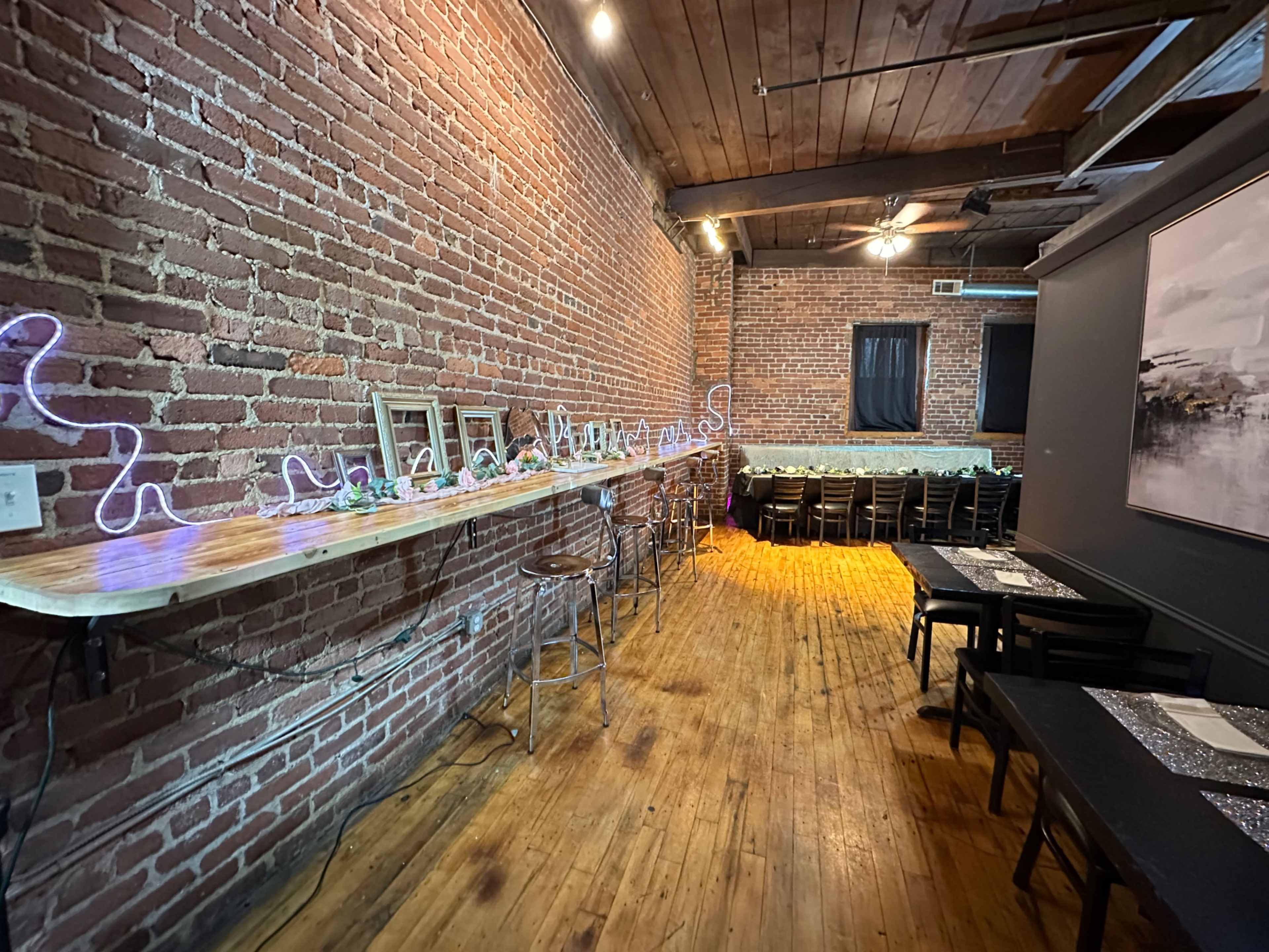 The image shows a long, narrow event space with exposed brick walls, wooden floors, a bar lined with seating, and tables set up for a gathering.