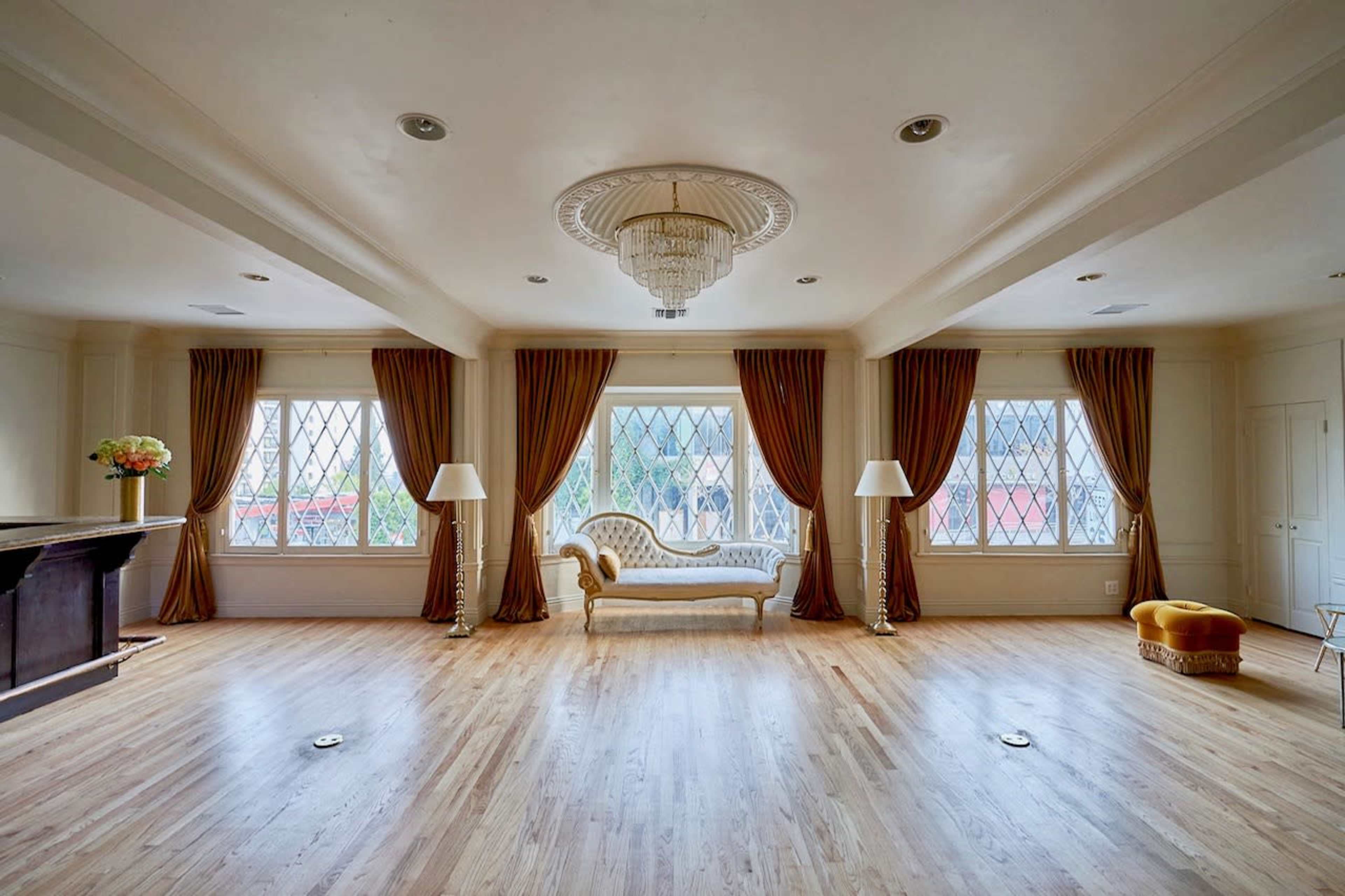 The room features a central chaise lounge flanked by two large windows draped in curtains, with a chandelier overhead and lamps at each end.