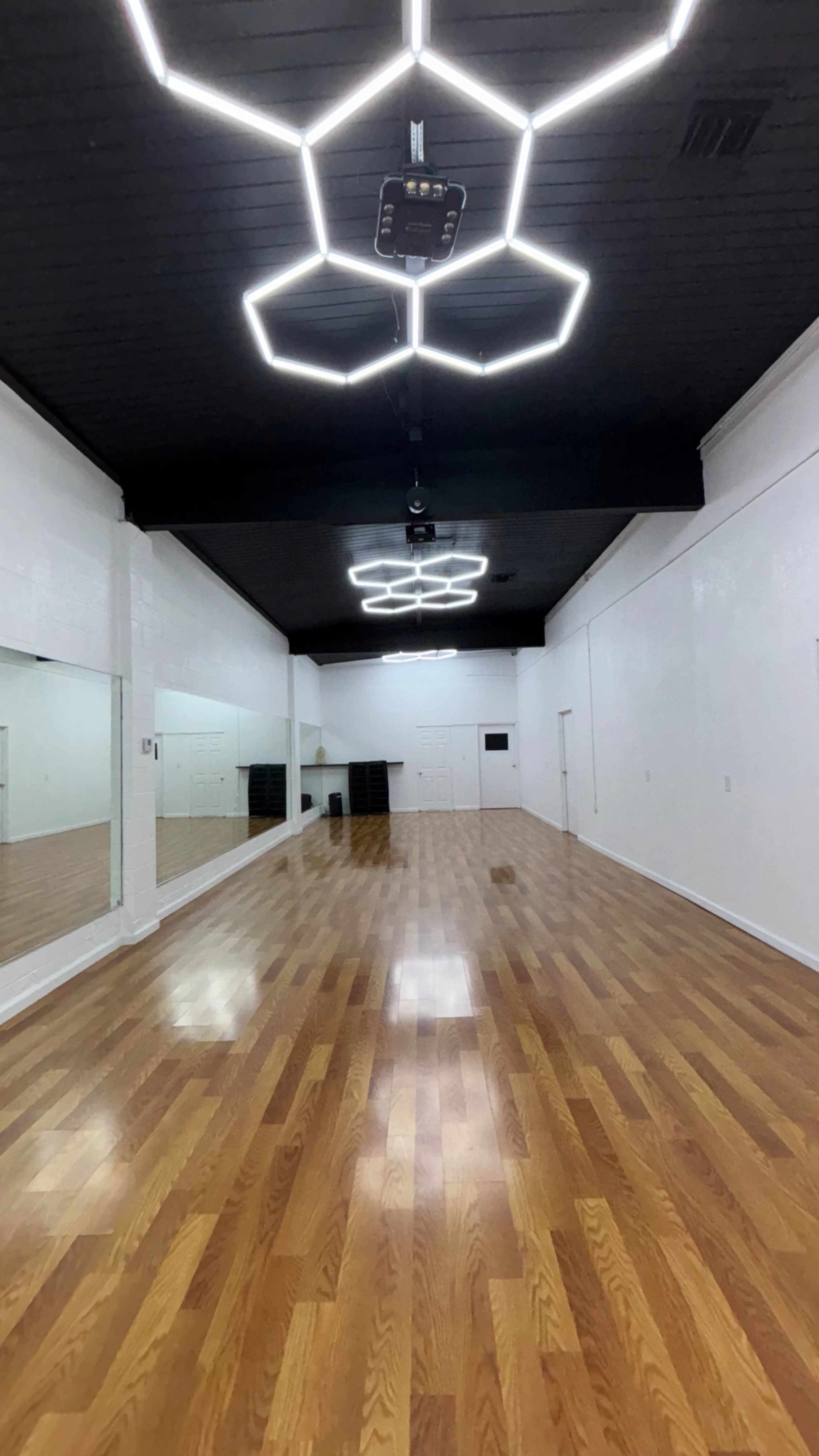 A spacious dance studio with wooden flooring, white walls, and hexagonal ceiling lights.
