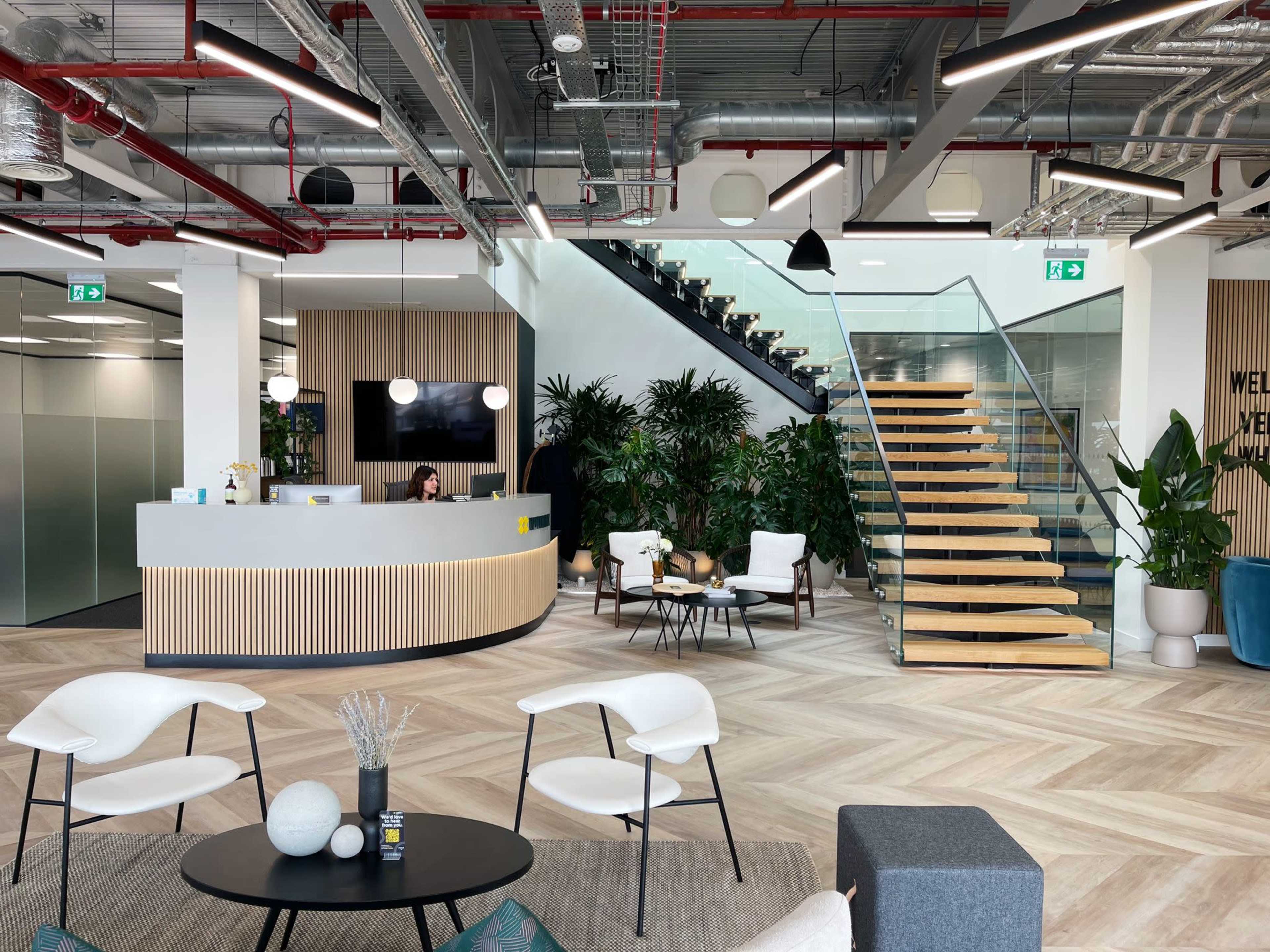 A modern office reception area with a curved desk, seating arrangements, greenery, and a prominent staircase in the background.