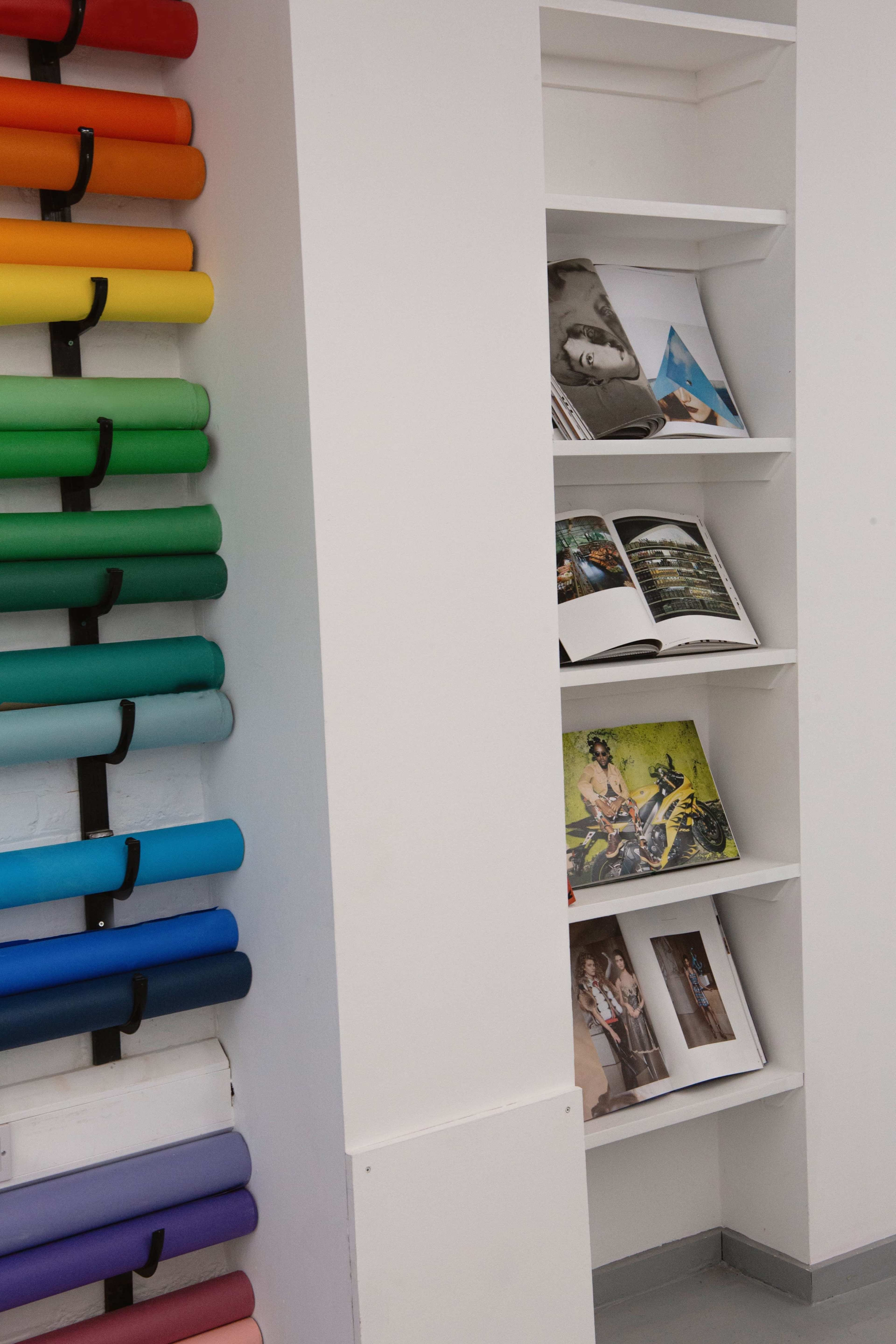 The image shows a wall with rolled paper in various colors on the left and a white shelf displaying magazines and photo books on the right.