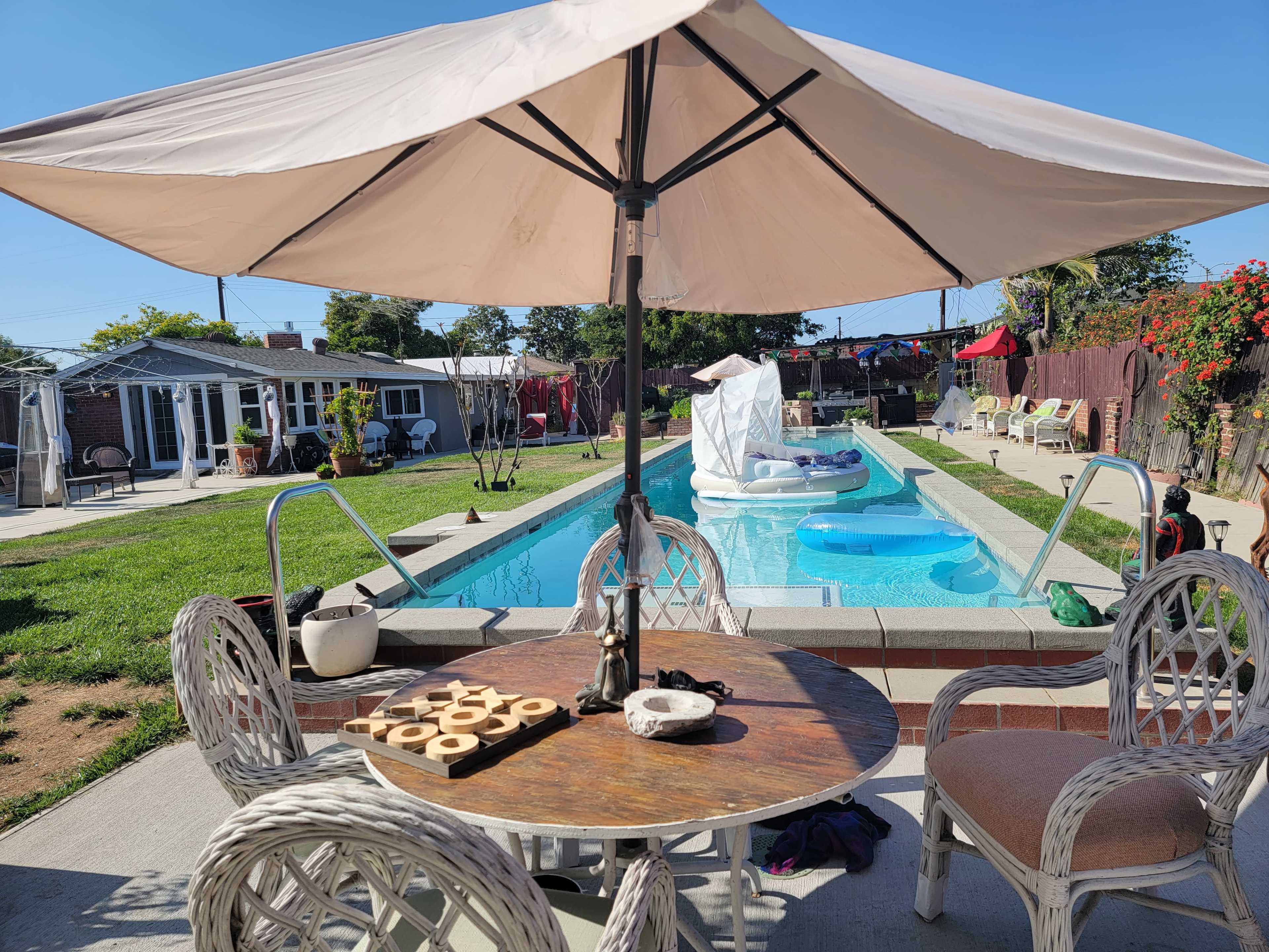 A backyard pool area with a table and chairs under an umbrella, a clean pool with inflatable toys, and various outdoor structures in the background.