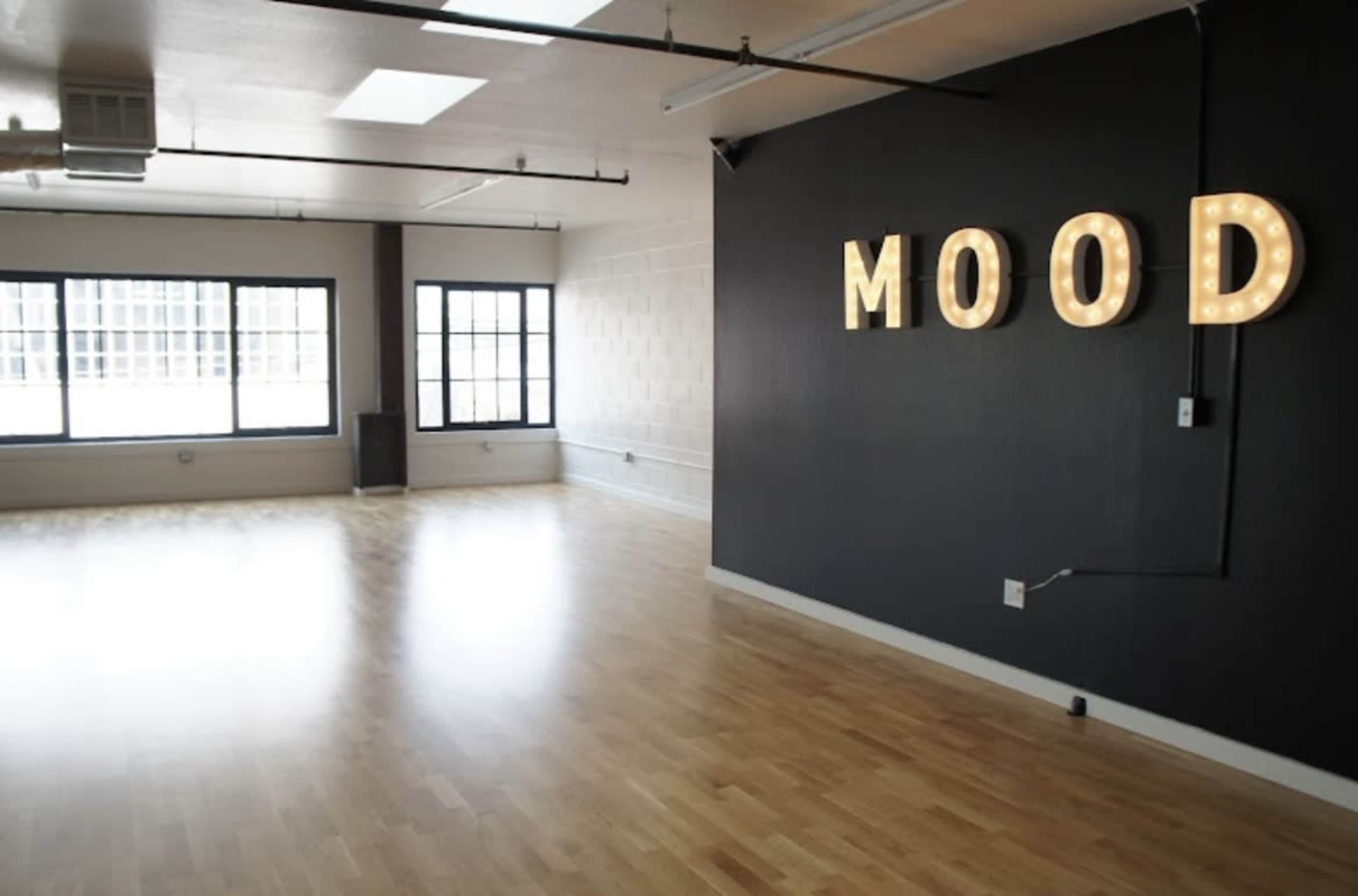 A spacious, modern room with wooden flooring and large windows, featuring illuminated letters that spell "MOOD" on one wall.