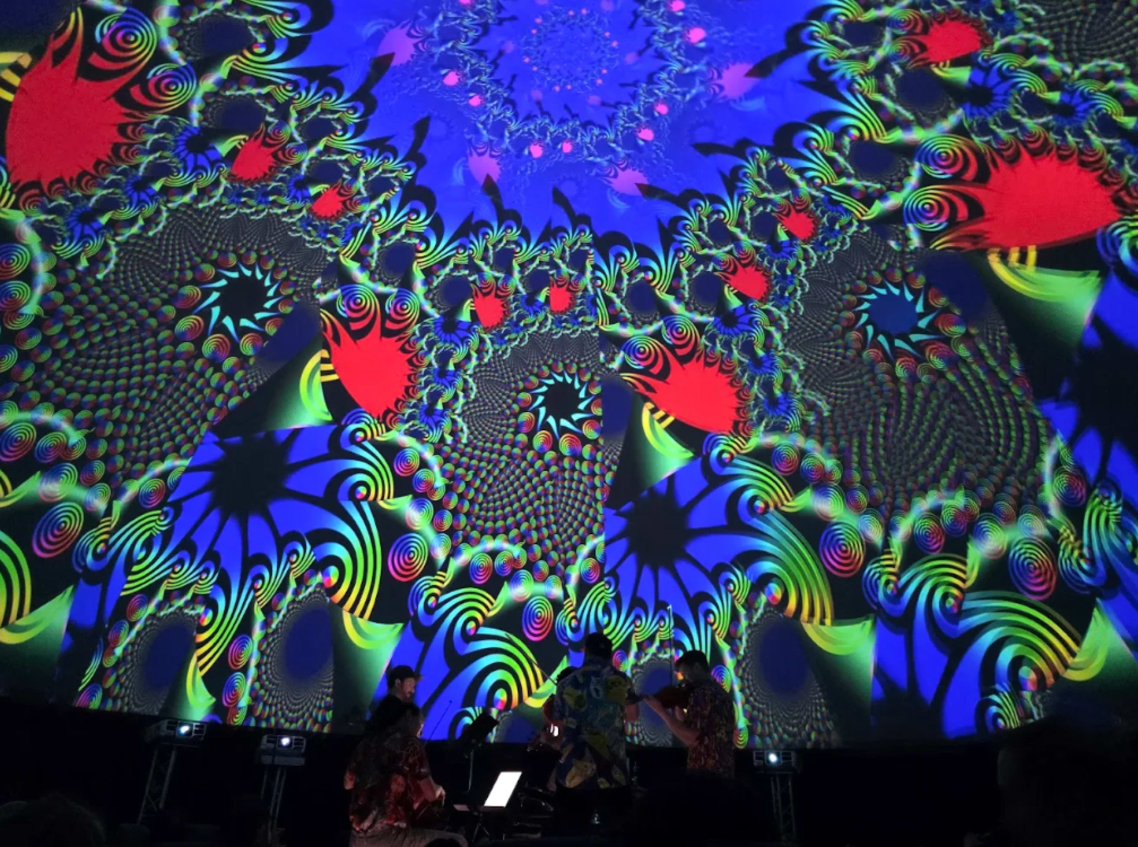 A group of musicians performs on stage beneath a vibrant, swirling psychedelic light display.