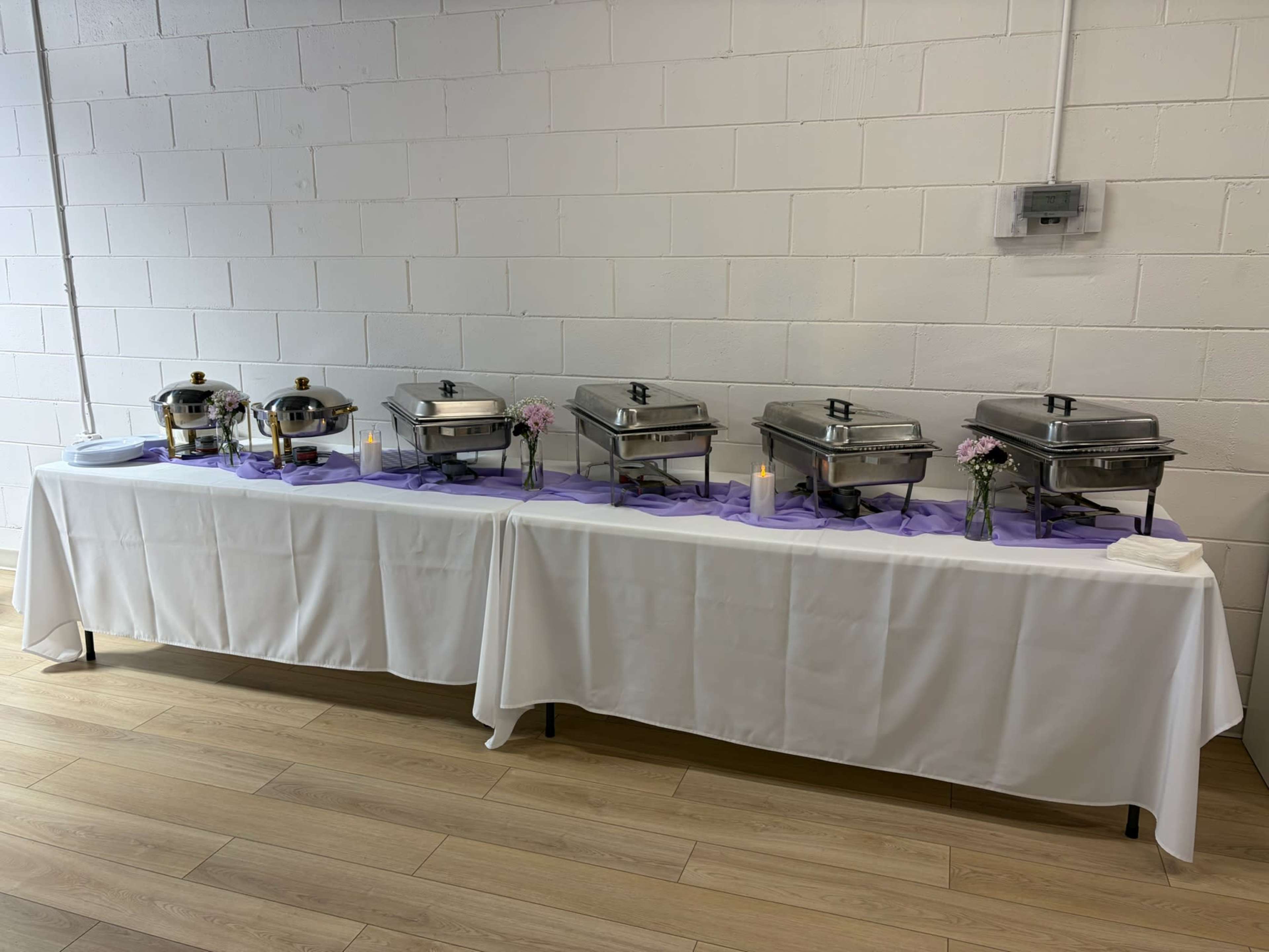 A row of chafing dishes is arranged on a table covered with a white cloth, with purple table runners and small vases of flowers placed between candles.