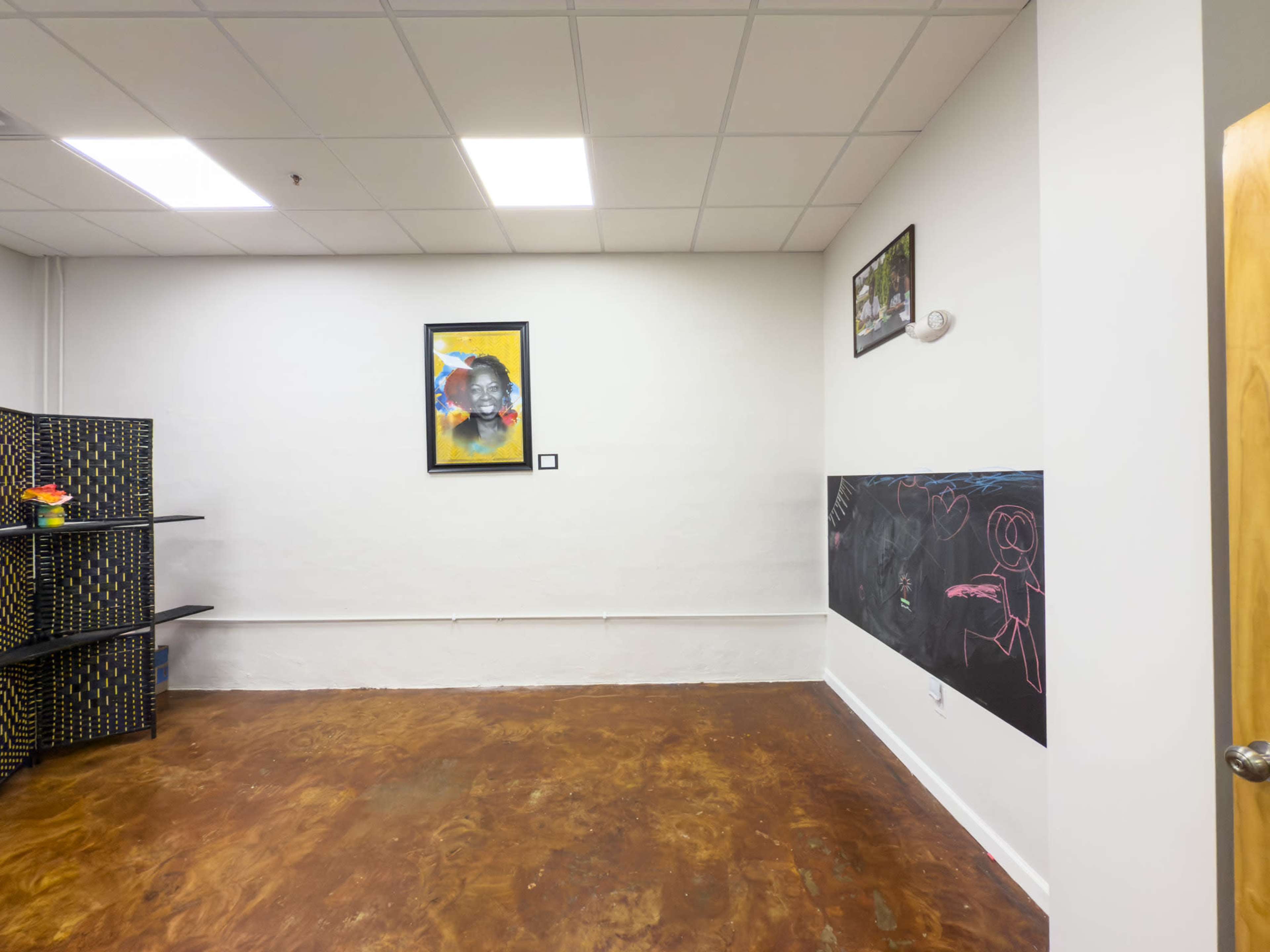 The image shows a small, brightly lit room with a painted concrete floor, a wall featuring framed artwork and a chalkboard, and a shelf filled with black boxes.