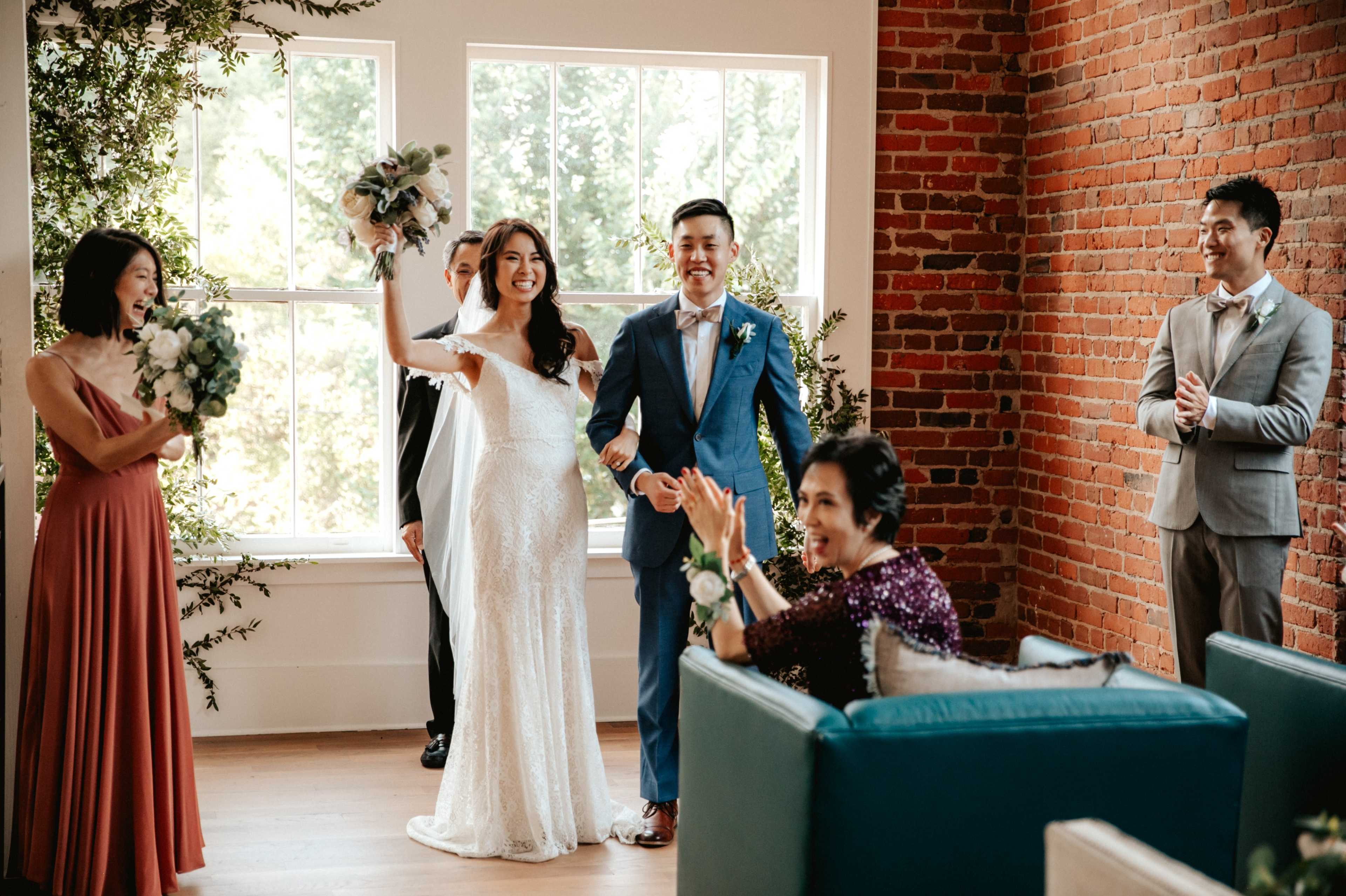A bride in a wedding gown and a groom in a suit stand together, smiling and celebrating with family and friends in a room with brick walls and large windows.