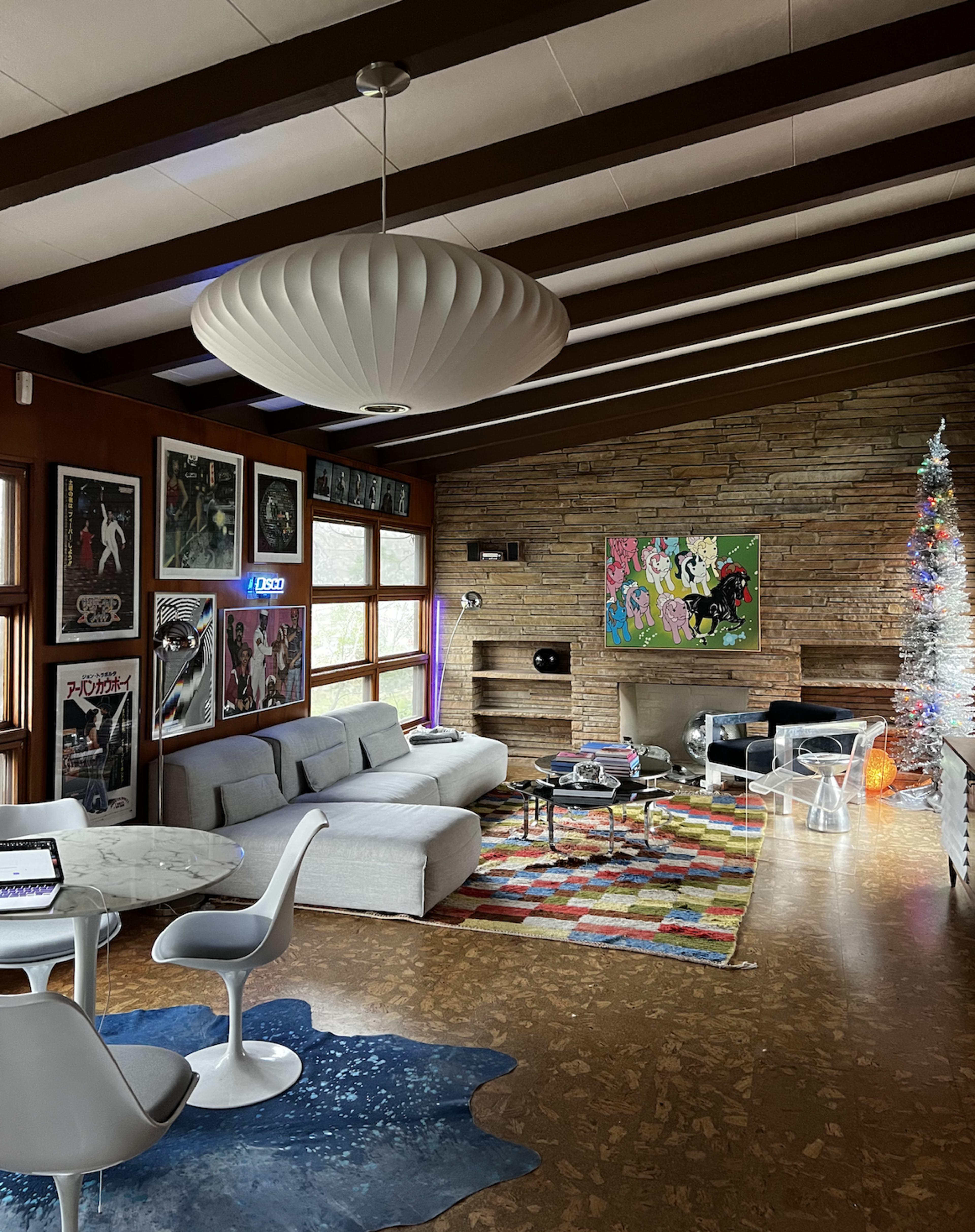 The room features a modern living area with a white sofa, colorful rug, and a decorated Christmas tree, alongside vintage posters on the walls and a stone fireplace.