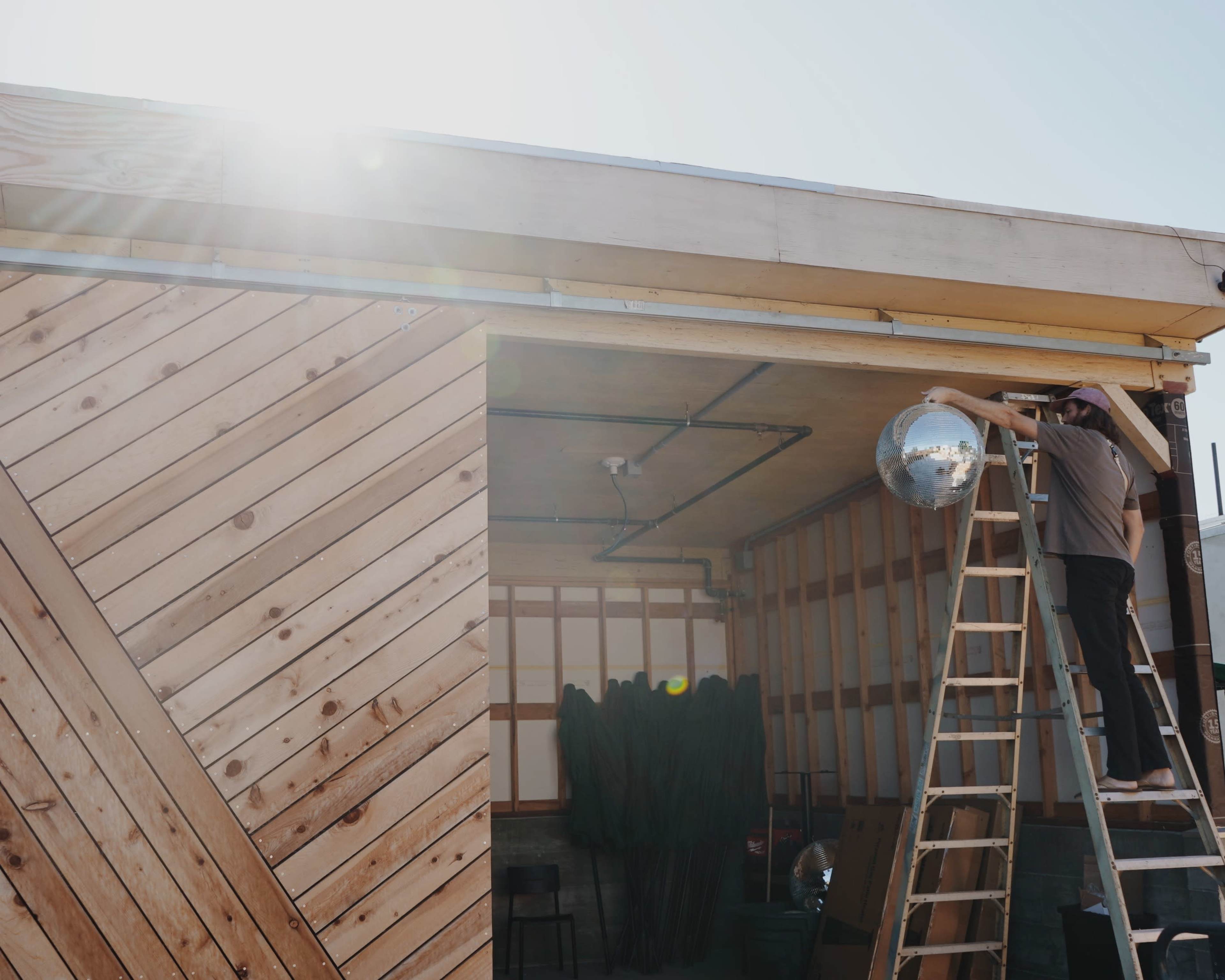 A person is climbing a ladder to hang a disco ball inside a wooden structure with diagonal paneling.
