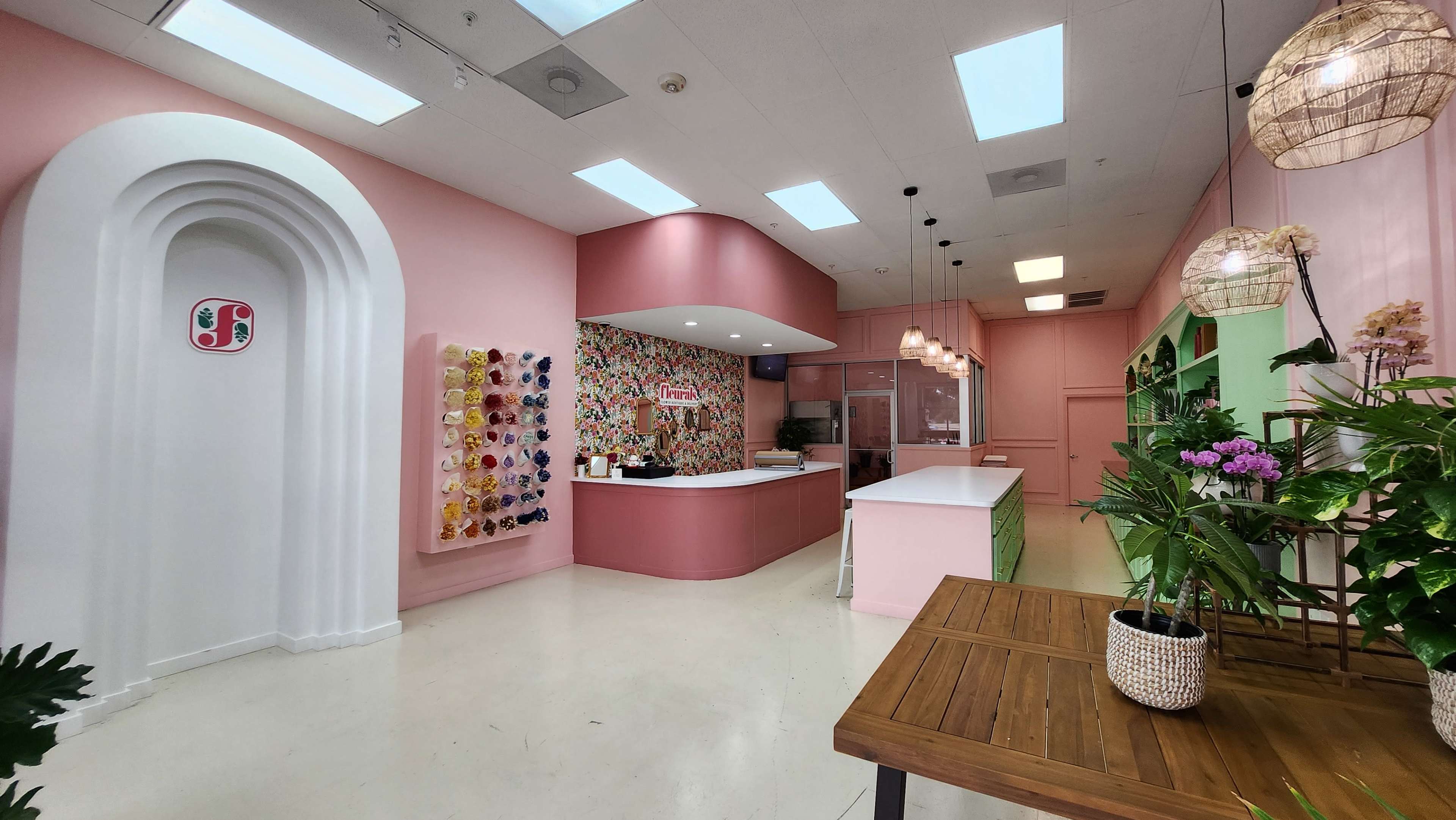 A brightly colored interior of a shop featuring a reception area with a pink counter, decorative wall art, and wooden tables with plants.