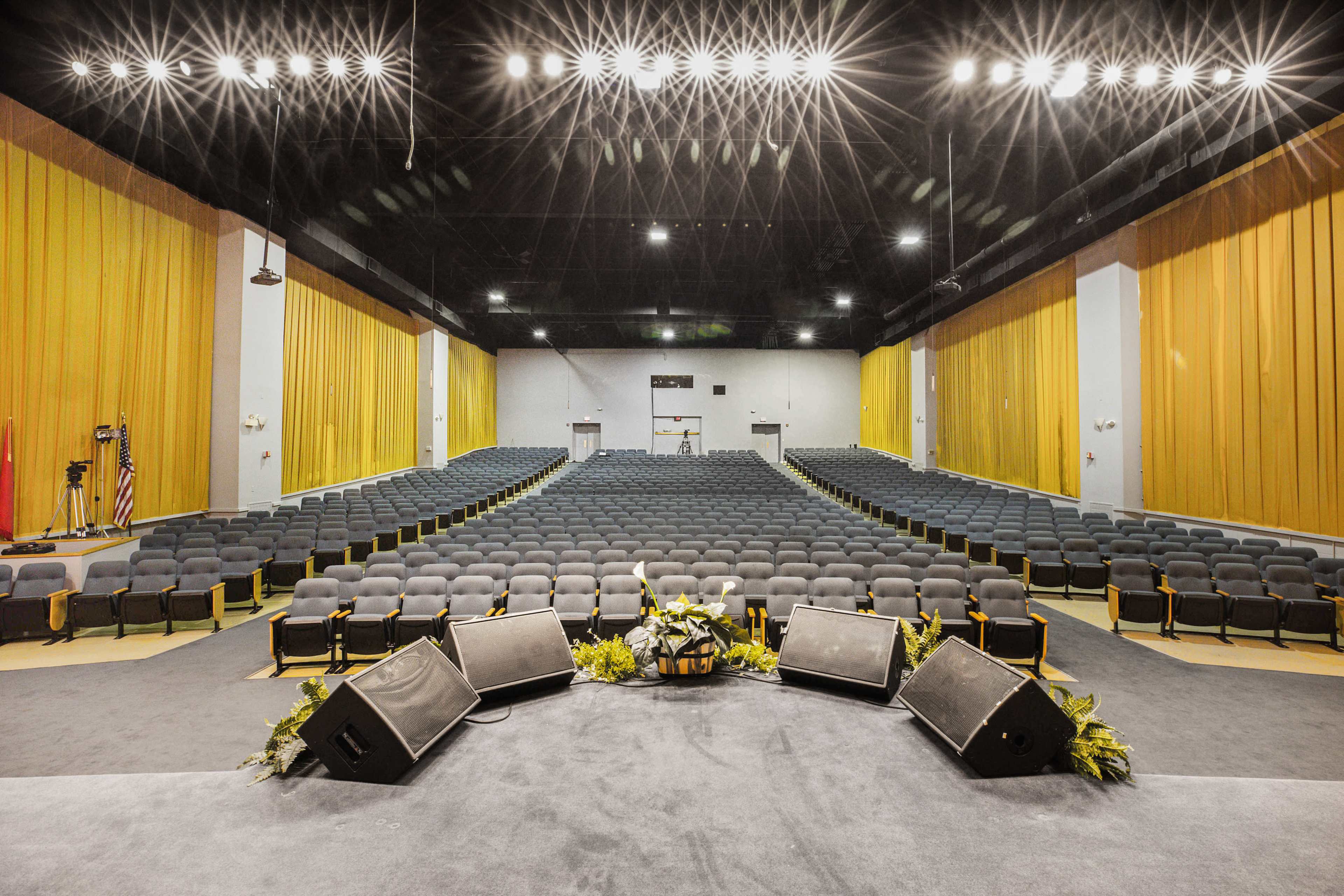 An empty auditorium with rows of black chairs and a stage at the forefront, surrounded by yellow curtains and brightly lit from above.