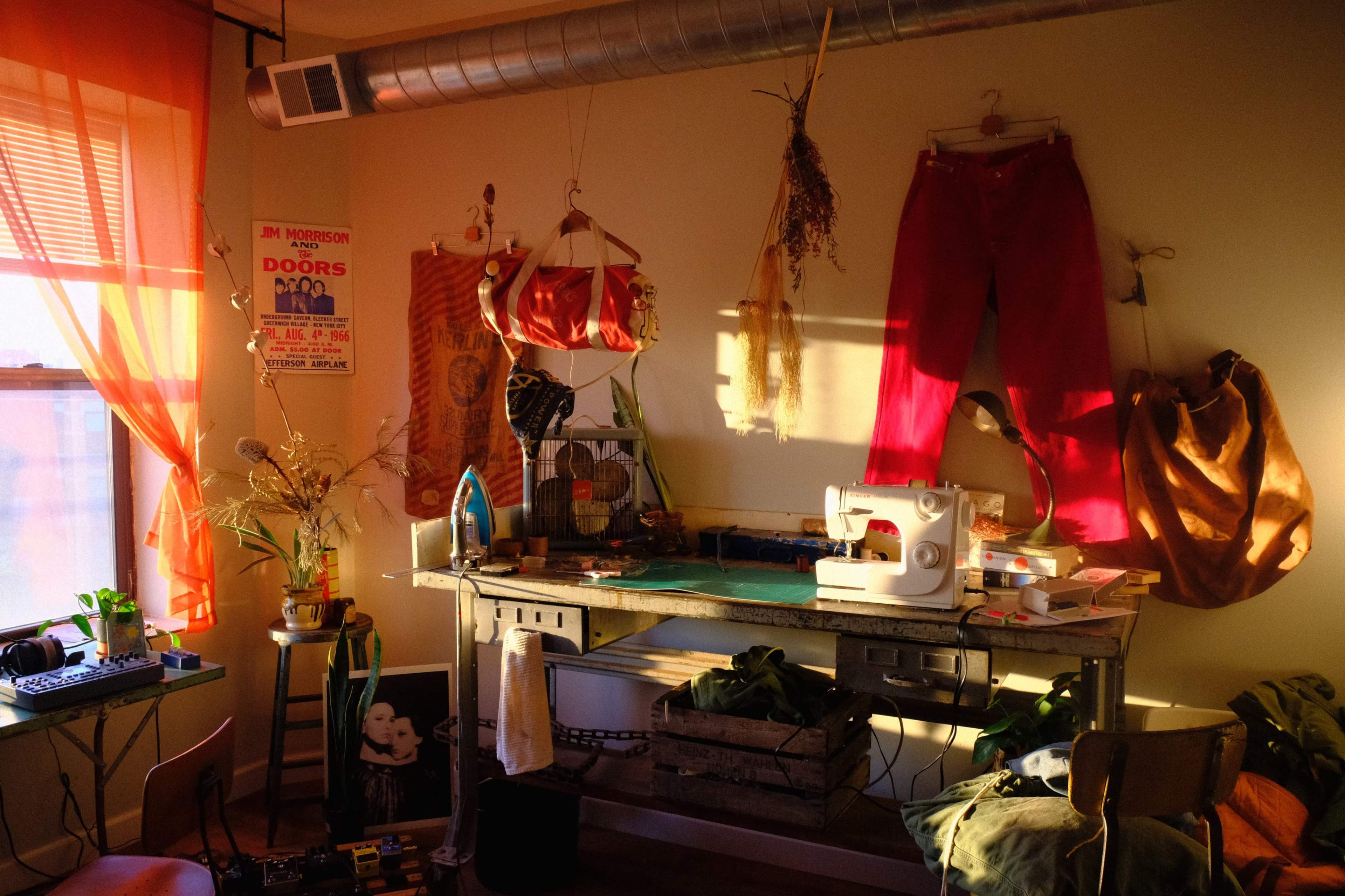 The image shows a brightly lit room featuring a sewing machine on a table surrounded by various sewing supplies, plants, and colorful curtains.