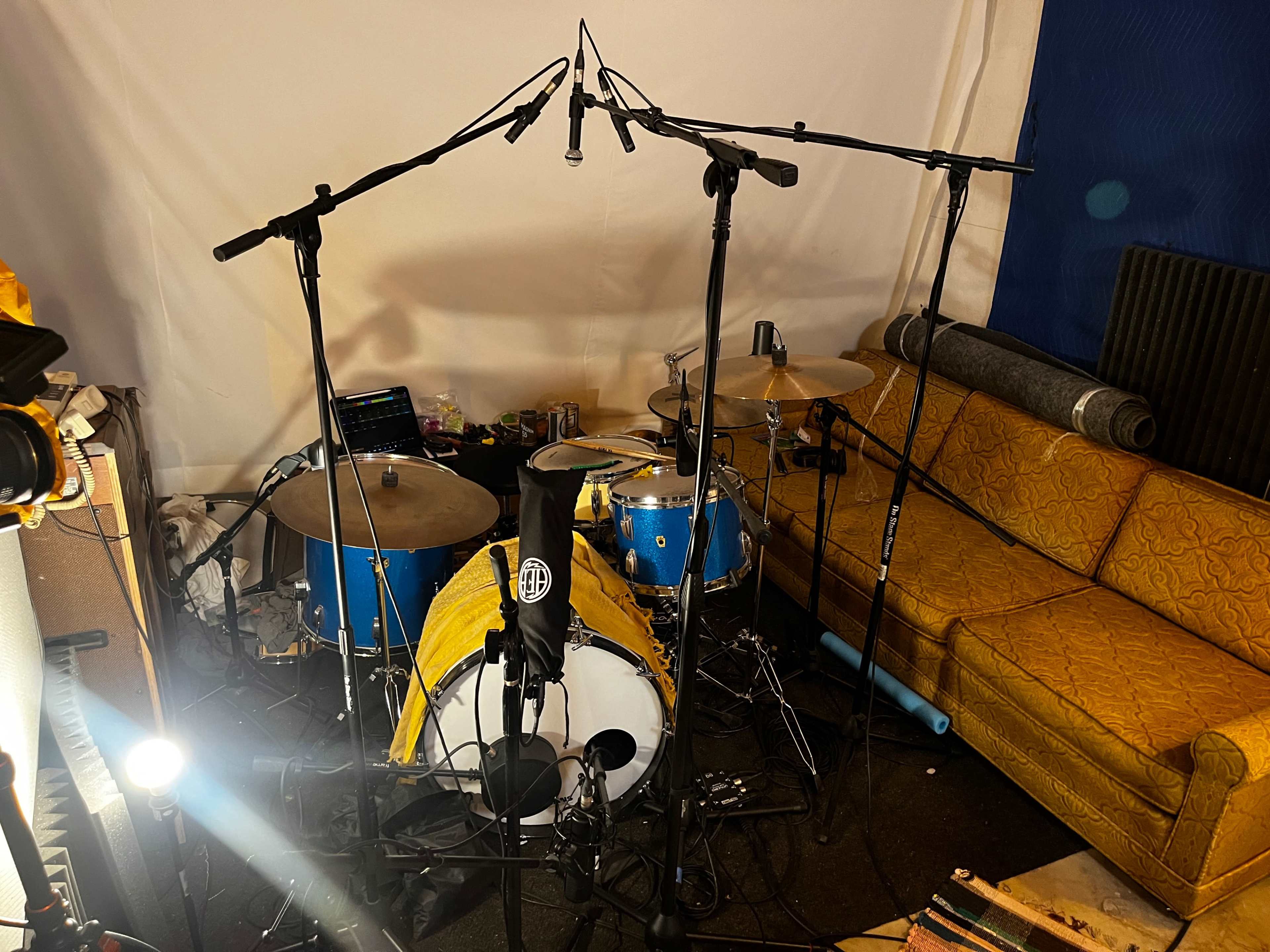 A small recording studio setup featuring a drum kit surrounded by microphone stands and a yellow couch in the background.