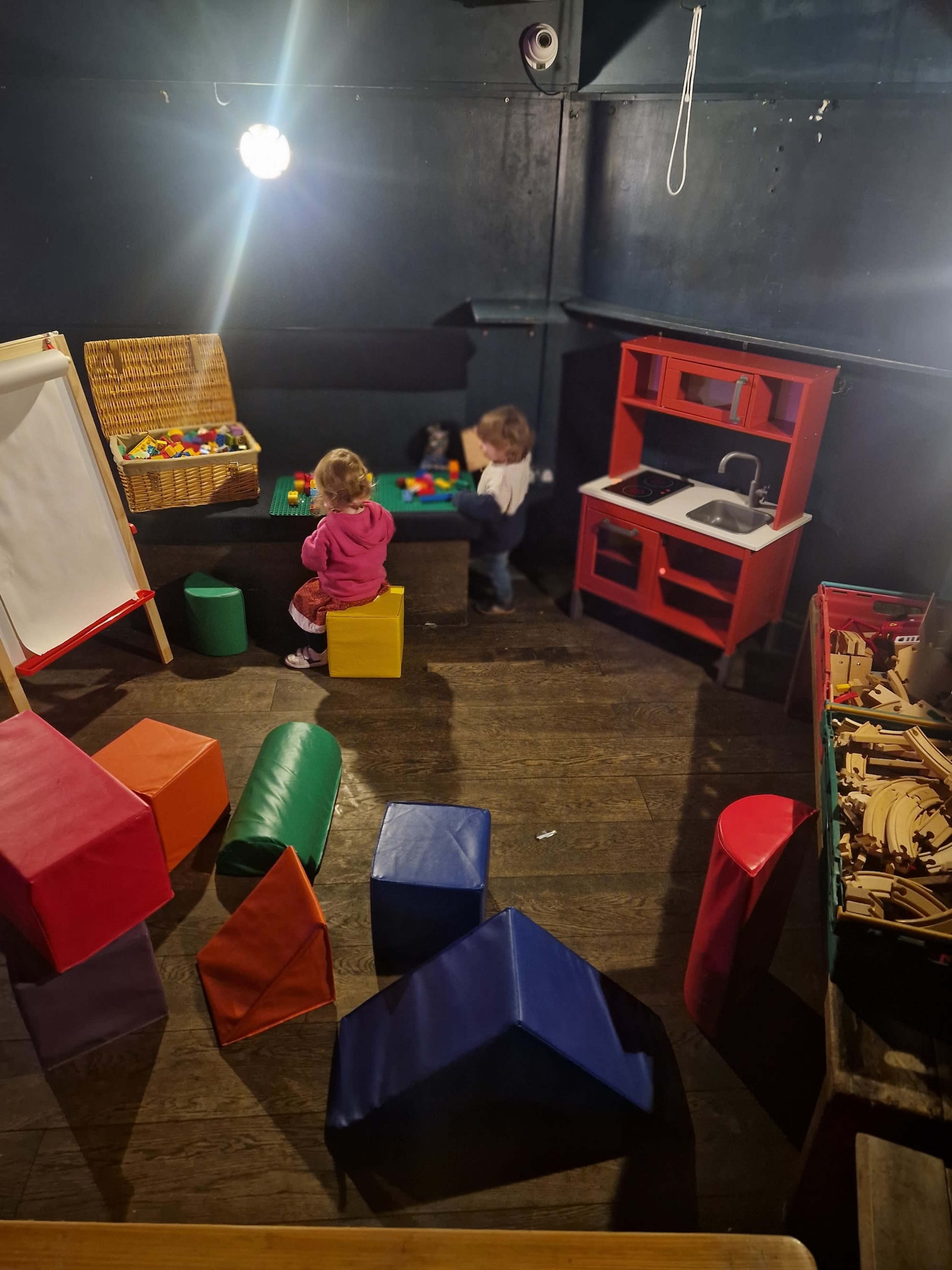 Two children are engaged in play at a small table surrounded by colorful geometric blocks in a room with dark walls and a play kitchen.