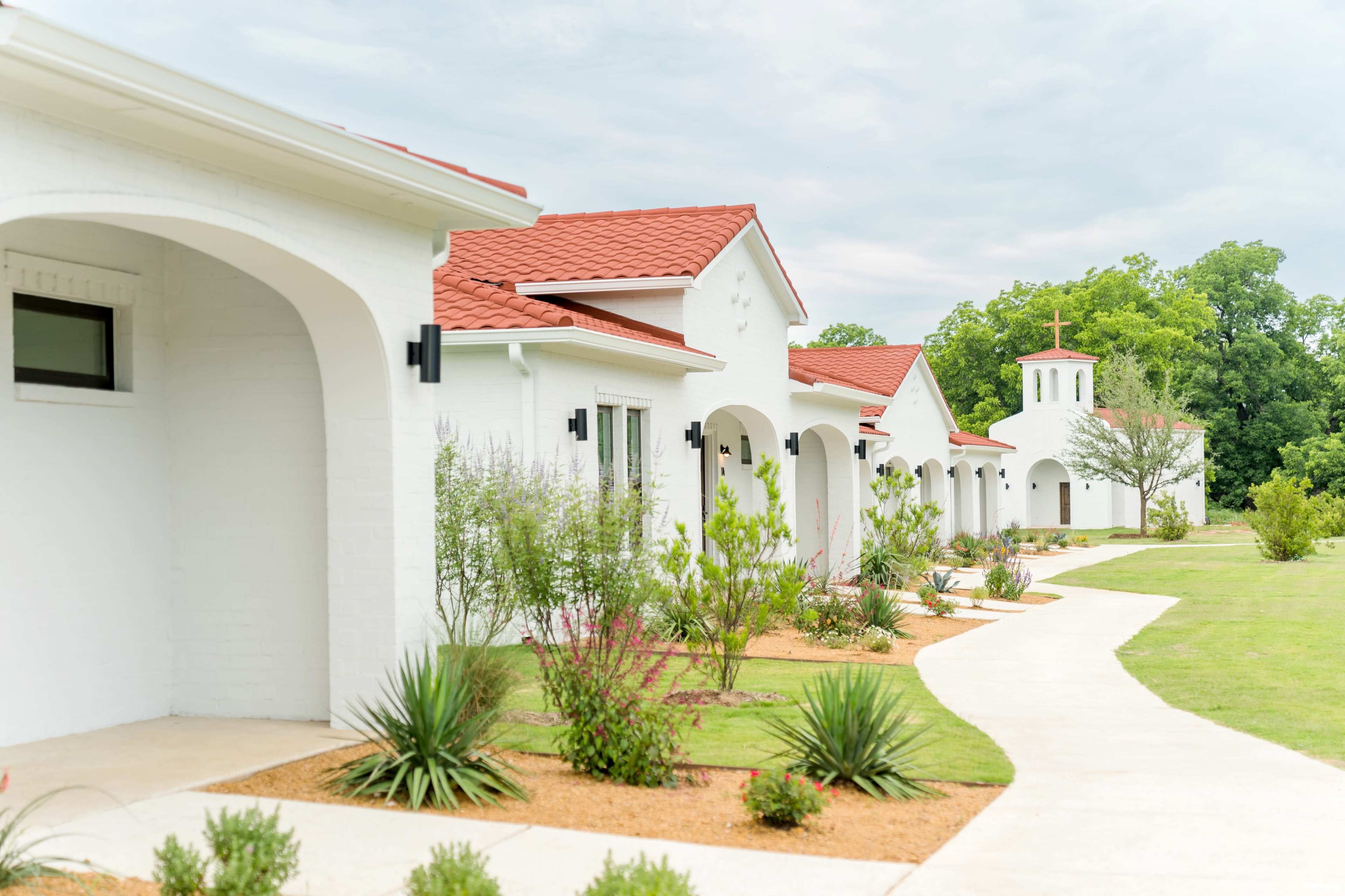 A row of white buildings with red roofs is lined along a pathway in a landscaped area with various plants.