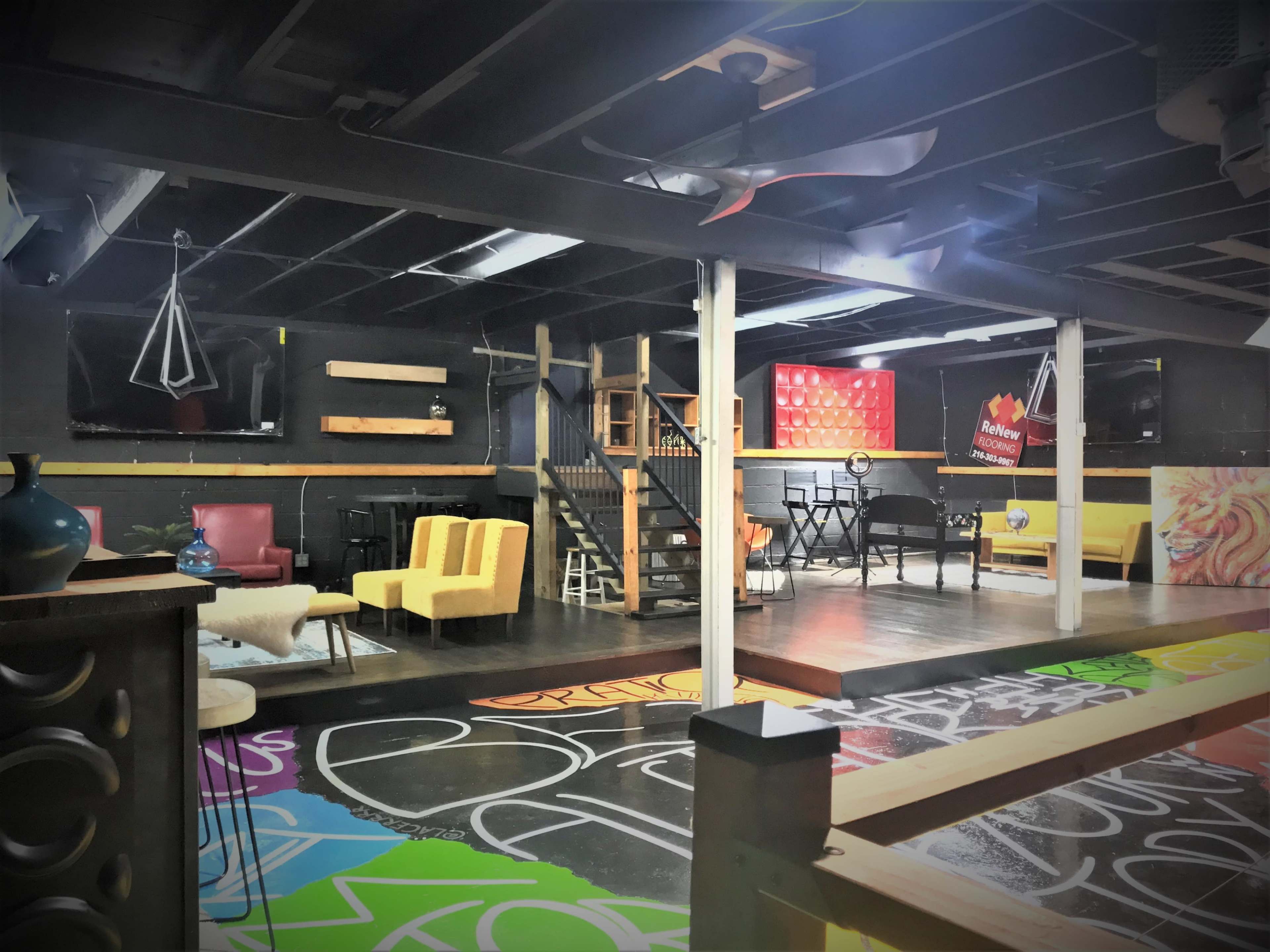 A spacious, colorful workshop or creative space featuring various seating areas, a staircase, and walls adorned with vibrant graffiti.