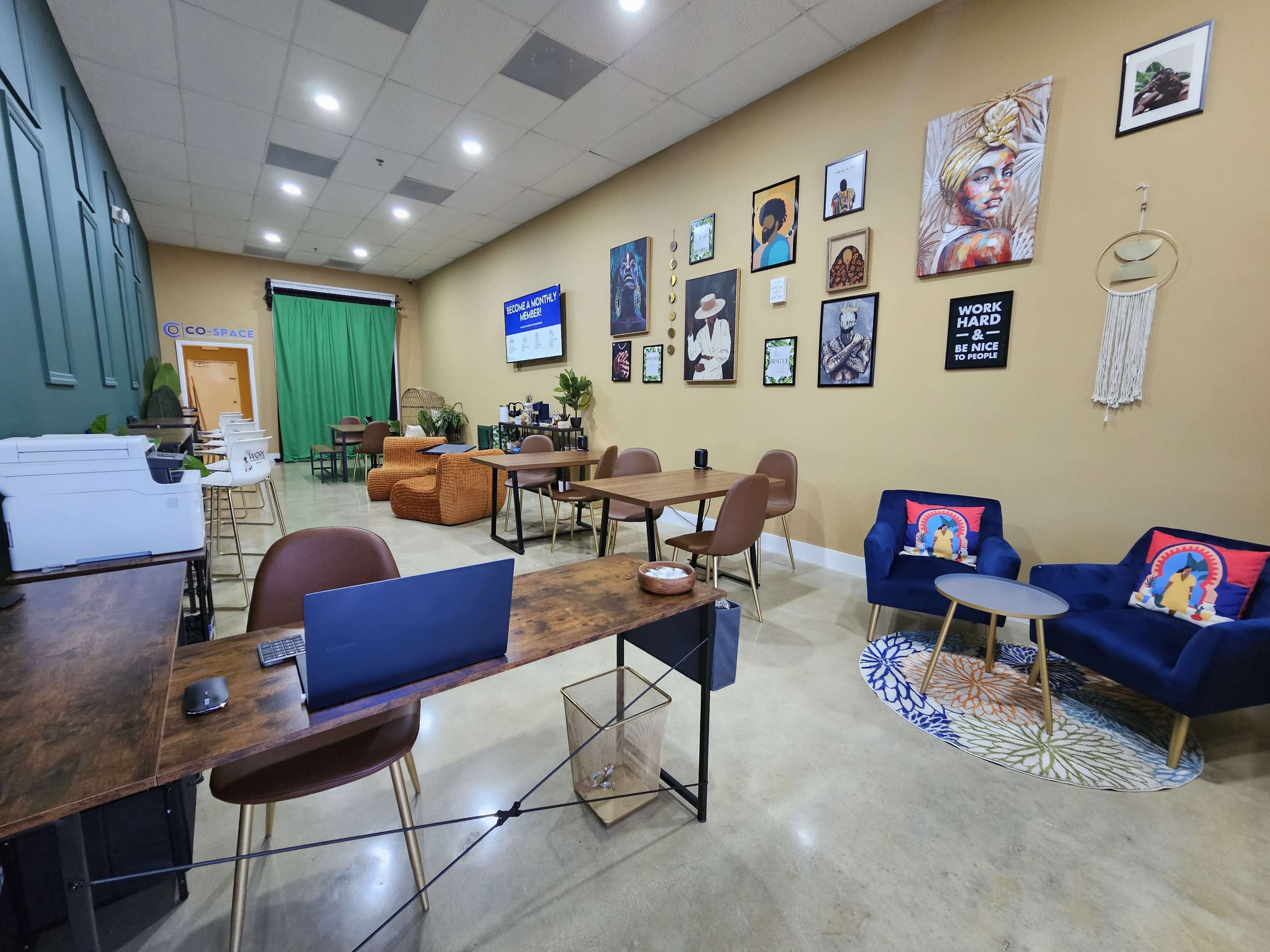 A co-working space with several tables, chairs, and a green screen backdrop, adorned with various artworks on the walls.