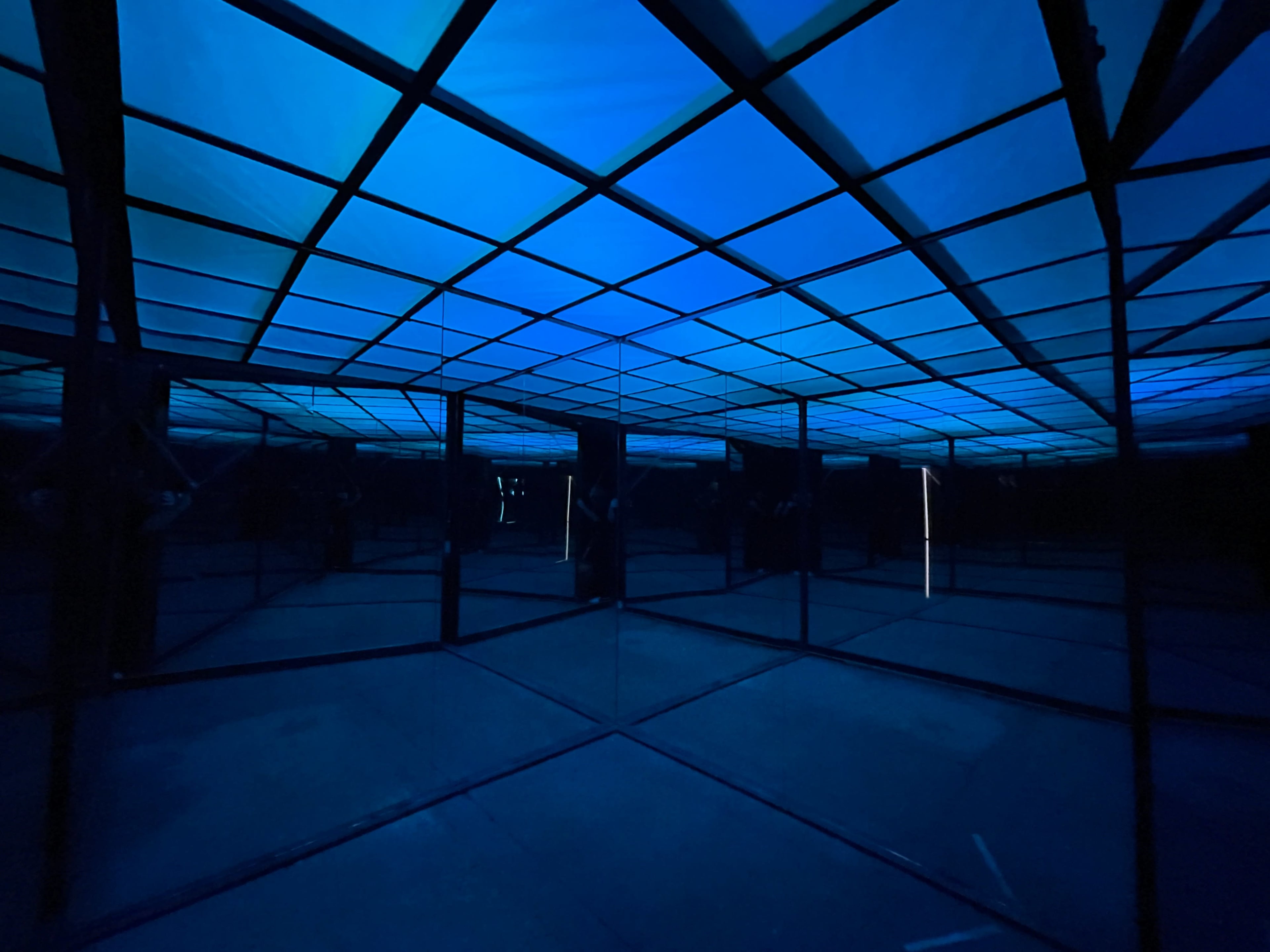 A dark room with a grid-patterned ceiling illuminated in blue, surrounded by mirrored walls creating a reflective environment.