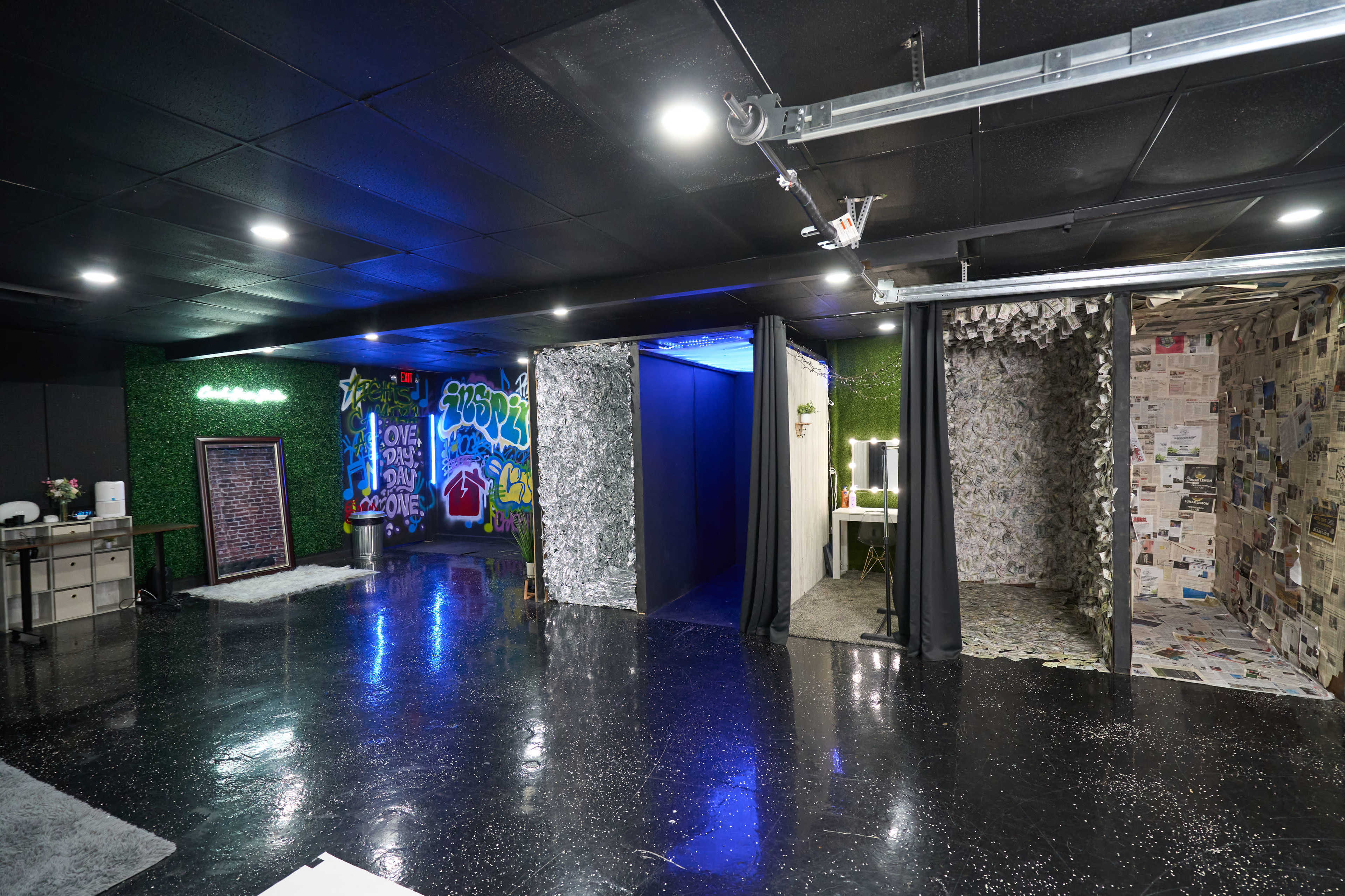The image shows a spacious room with varying wall textures, including graffiti, fabric drapes, and newspaper decor, alongside illuminated mirrors and a well-organized workspace.