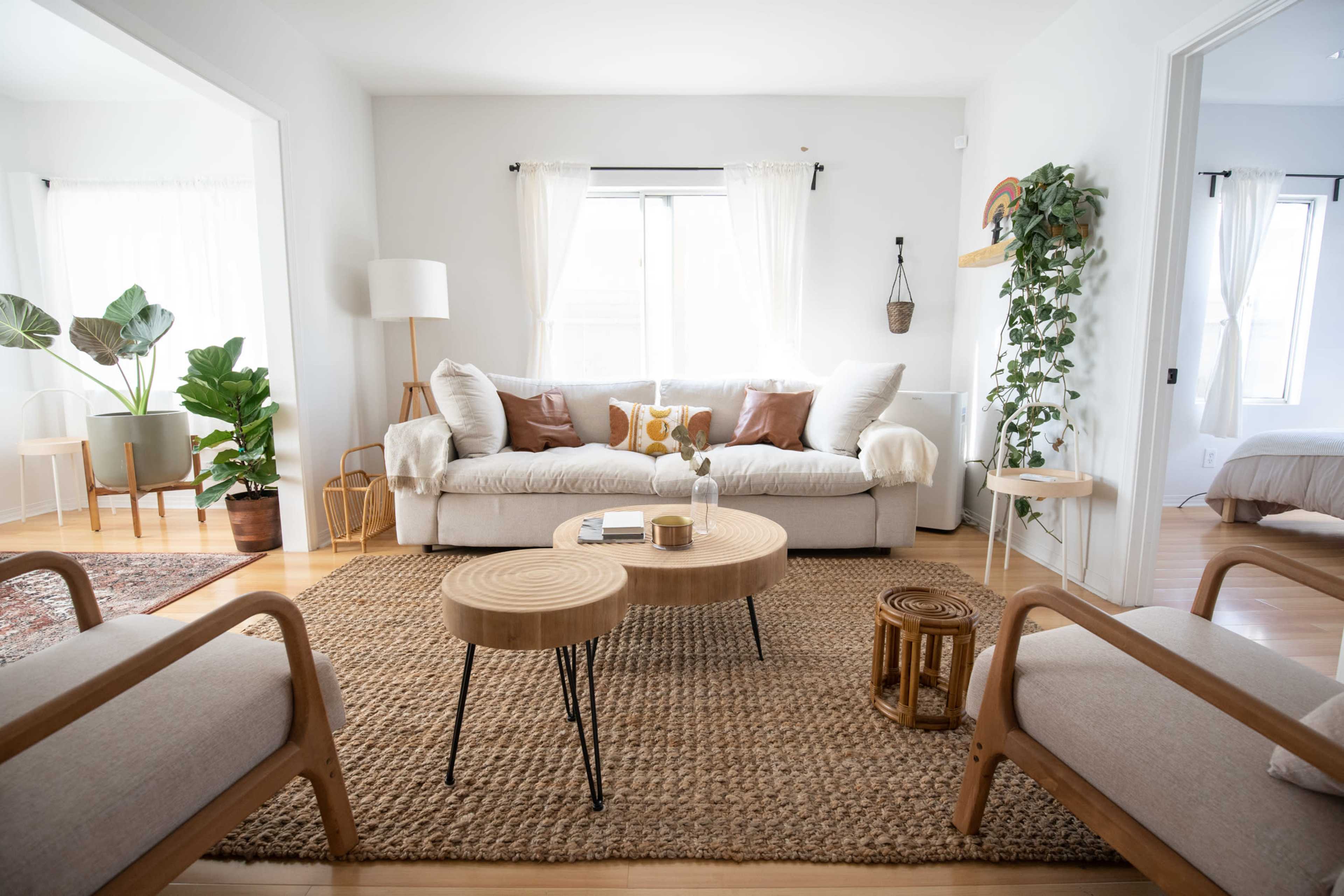 A bright living room featuring a beige couch with decorative pillows, two wooden chairs, a woven rug, and a pair of coffee tables.