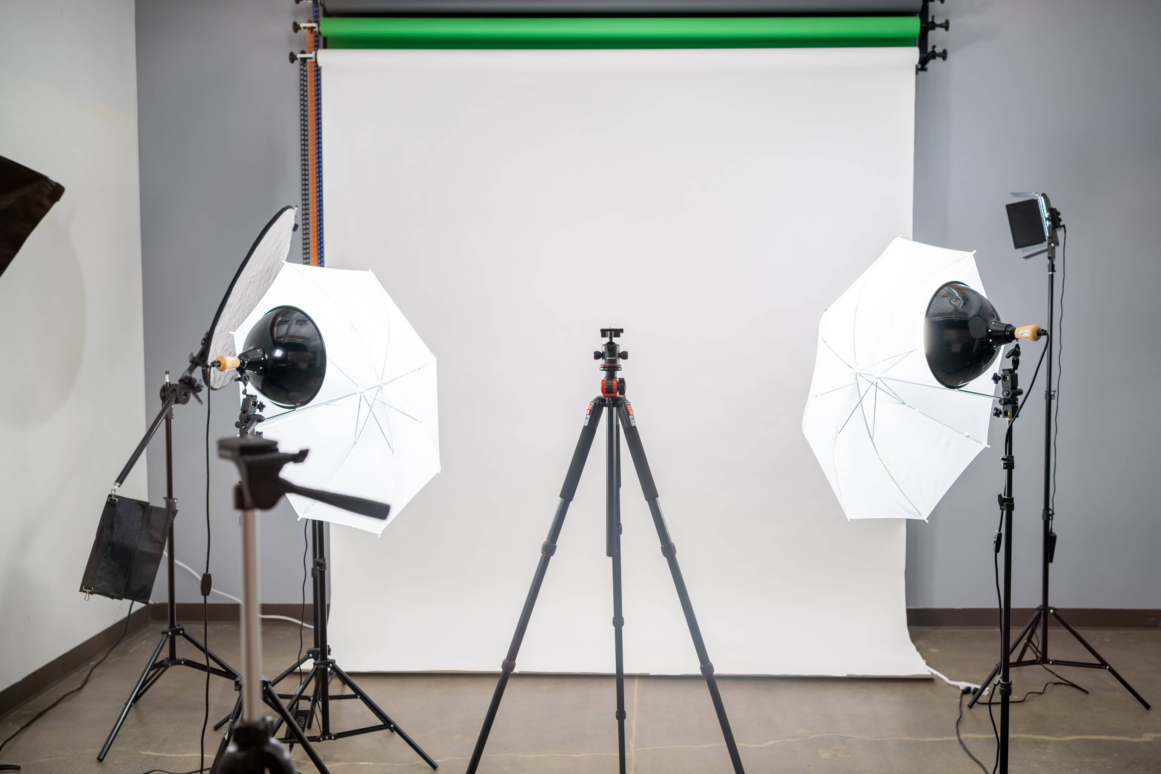A photography studio setup with two softbox lights, a tripod in the center, and a plain backdrop.