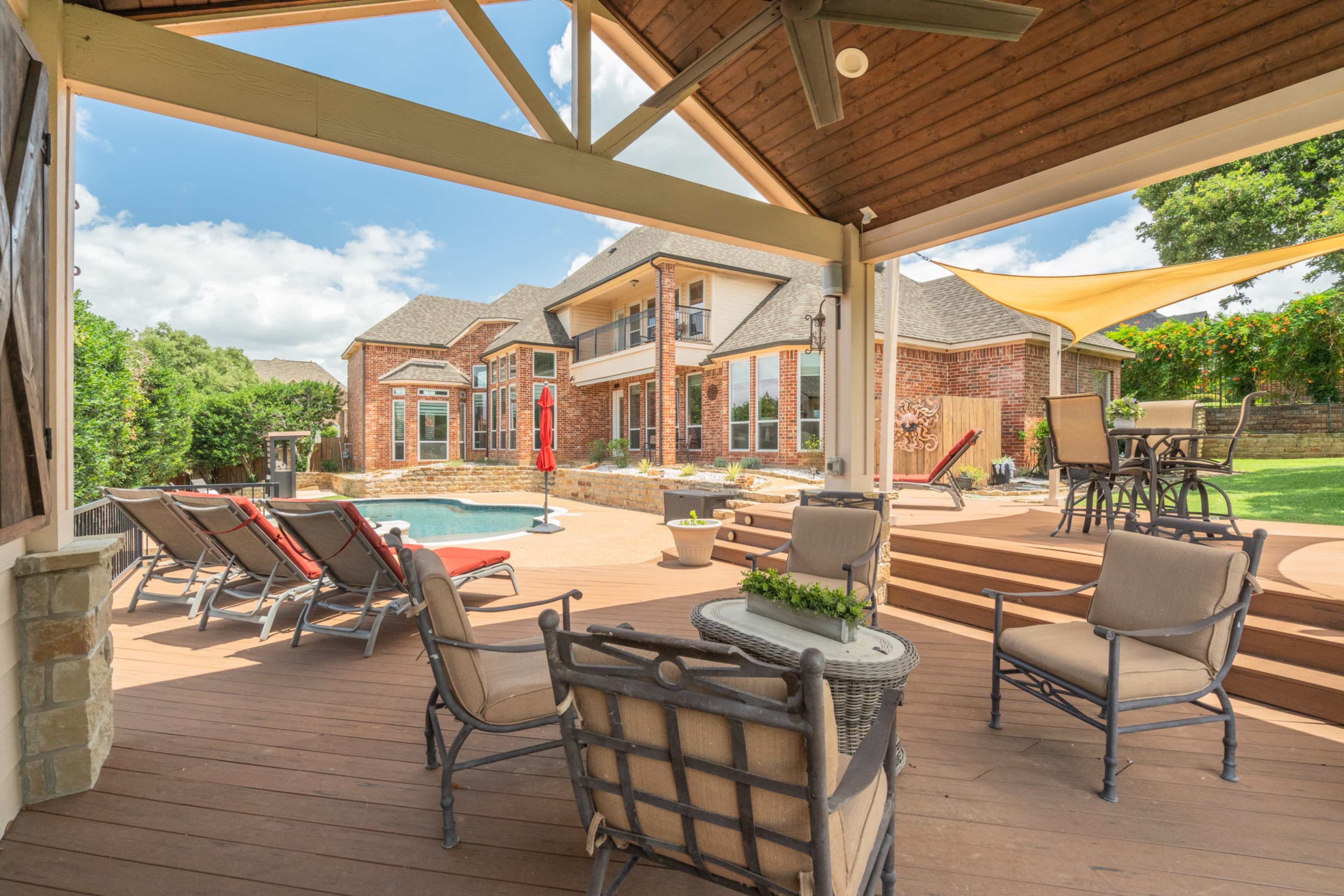 The image shows a backyard patio with seating arrangements overlooking a swimming pool and a two-story brick house in a residential setting.