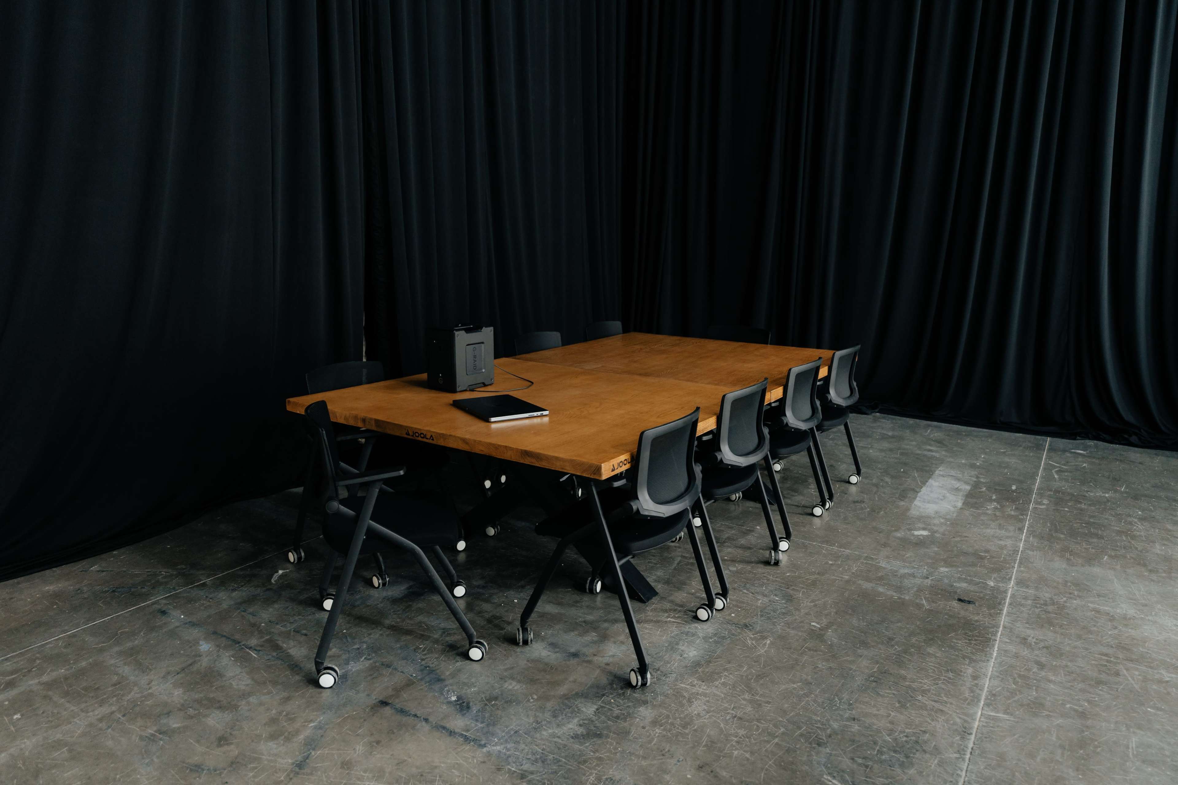 A large wooden table surrounded by several black chairs is set against a backdrop of dark curtains in a spacious room.