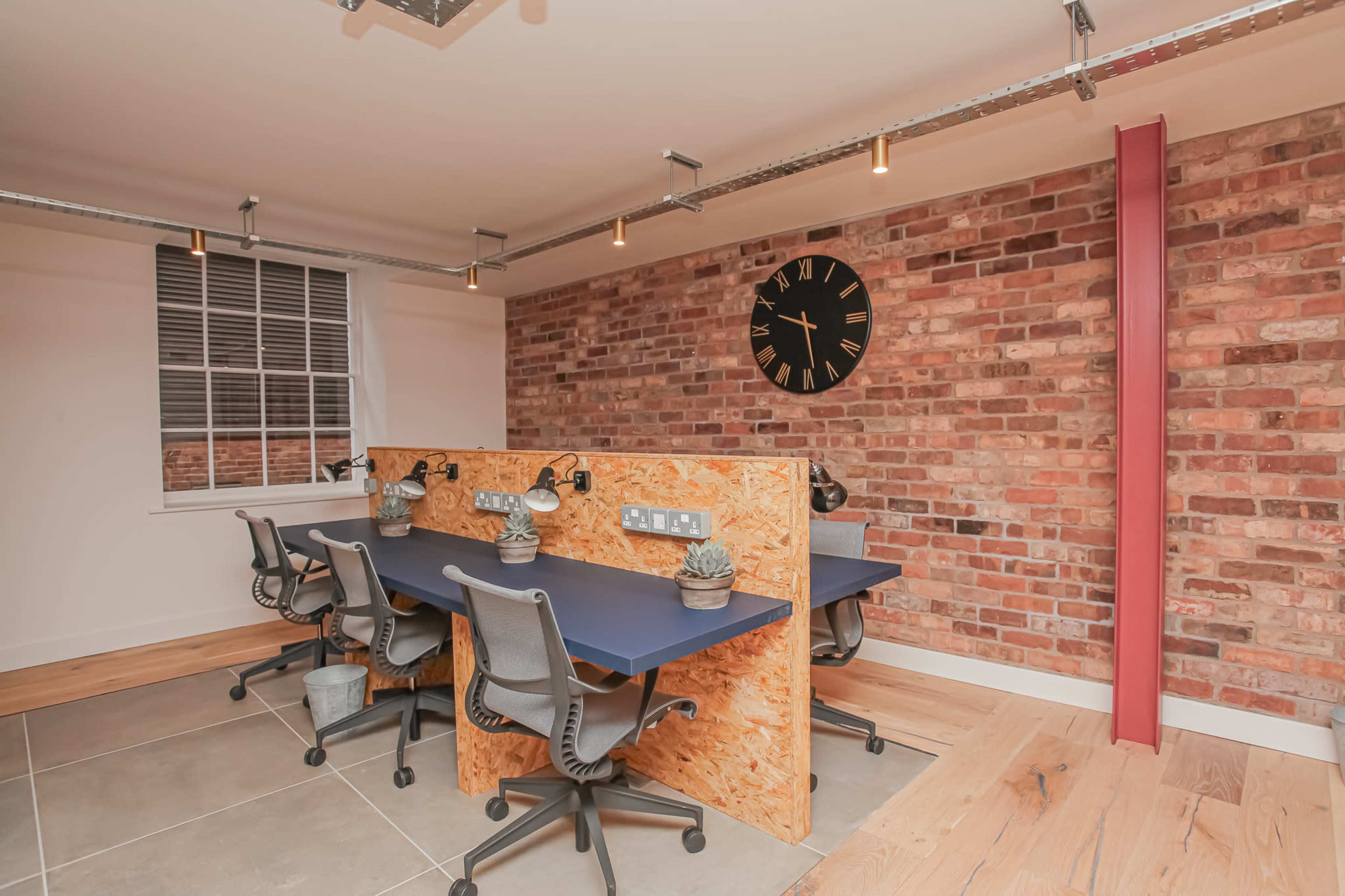 The image shows a modern office space with four workstations separated by wooden dividers, a brick wall in the background, and a clock mounted on the wall.