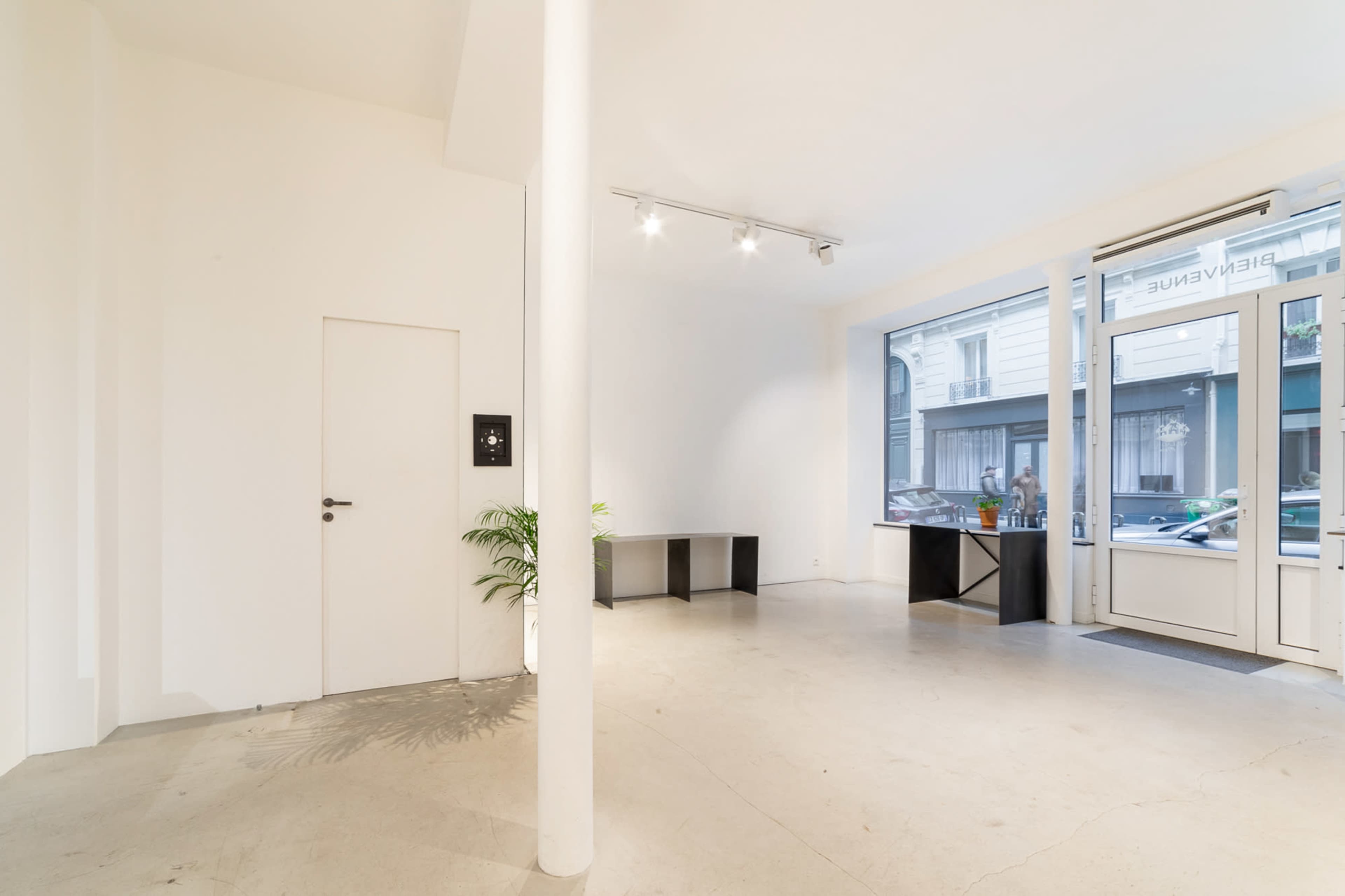 A minimalist interior of a white-walled gallery with a door, a simple table, and large windows revealing street views.