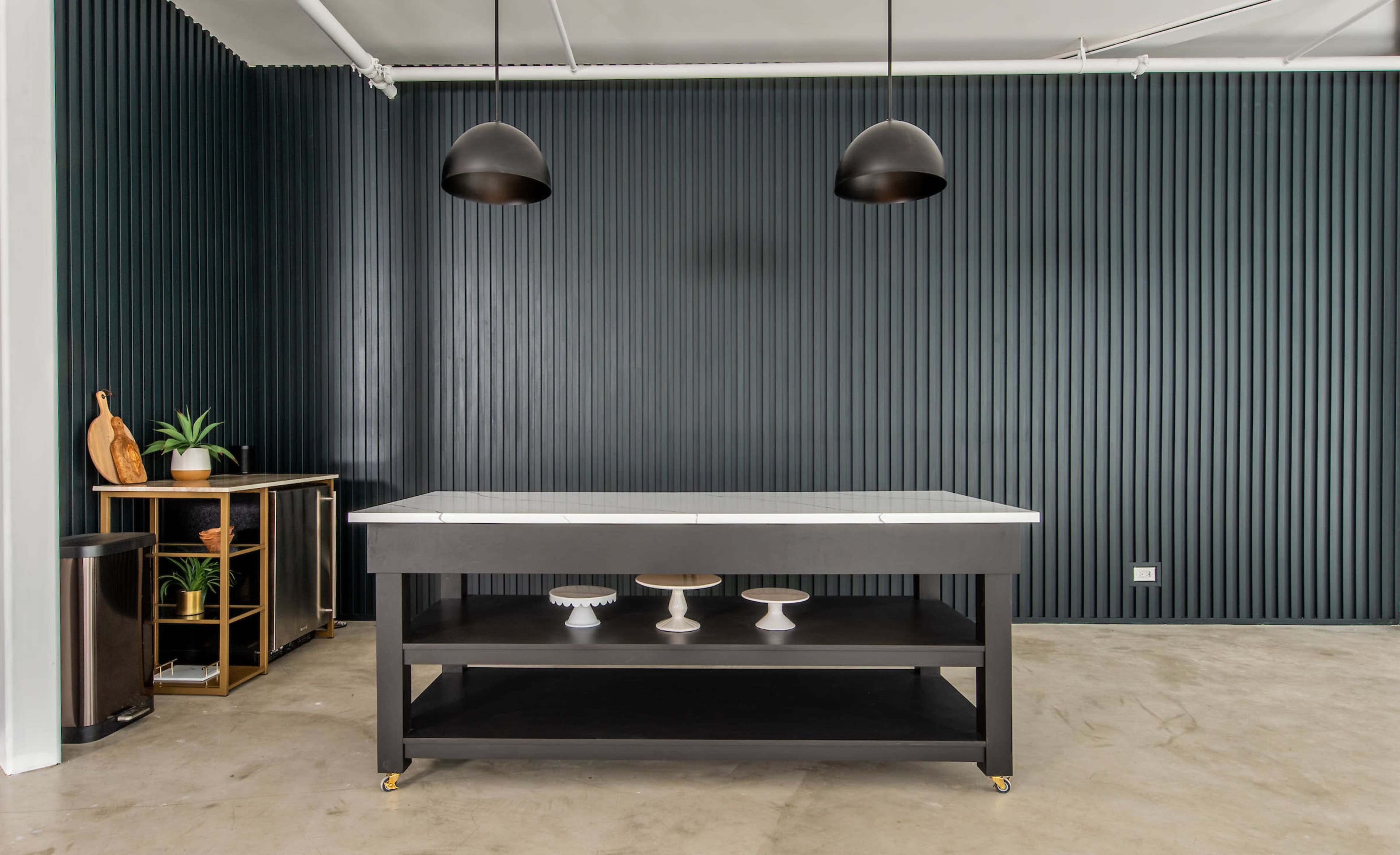 A black kitchen island with a marble top is positioned against a dark ribbed wall, accompanied by two hanging pendant lights and a wooden table displaying ceramics.