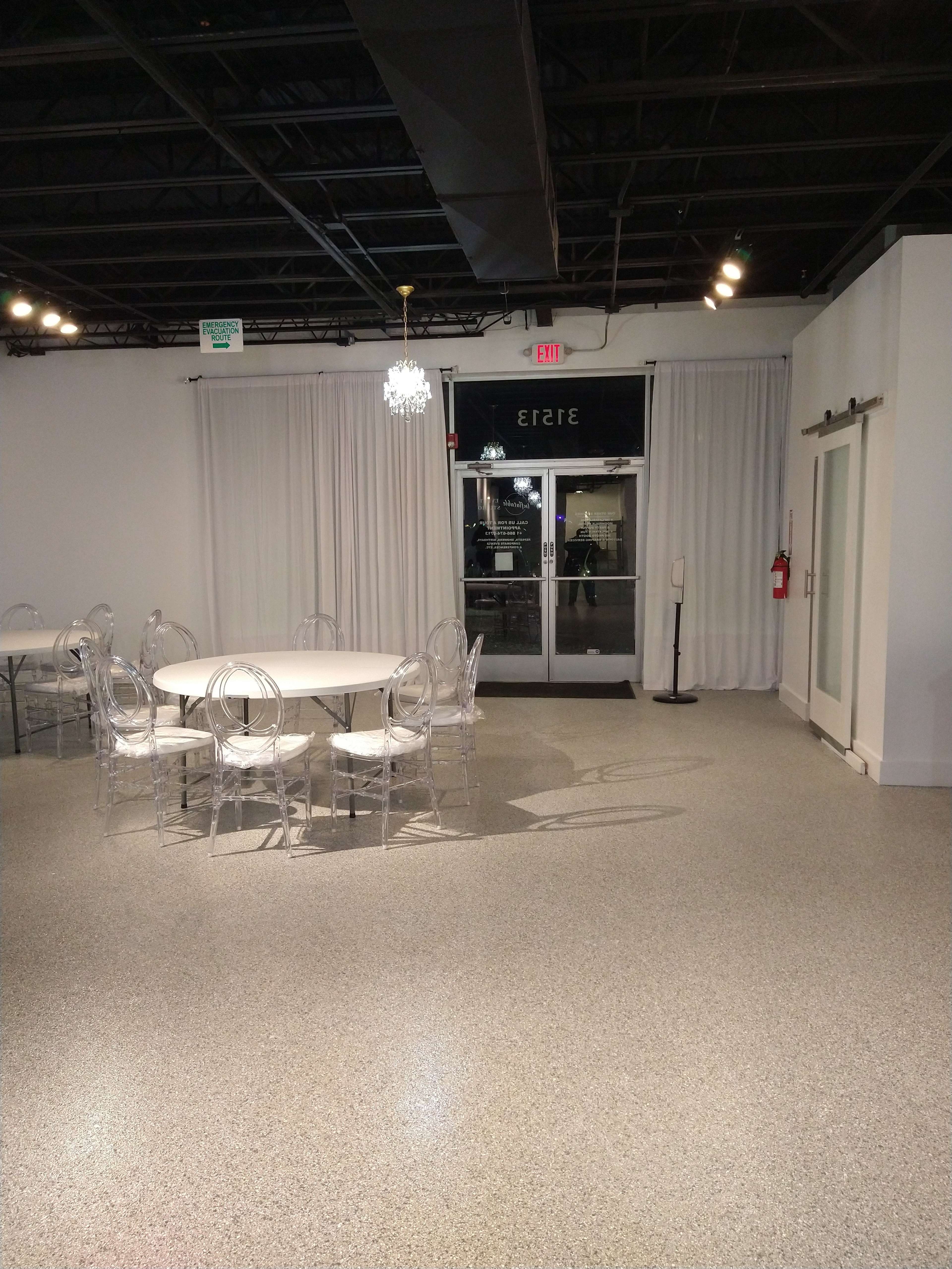The image shows a spacious, empty event room with a round table surrounded by clear chairs, white curtains, and exit doors.