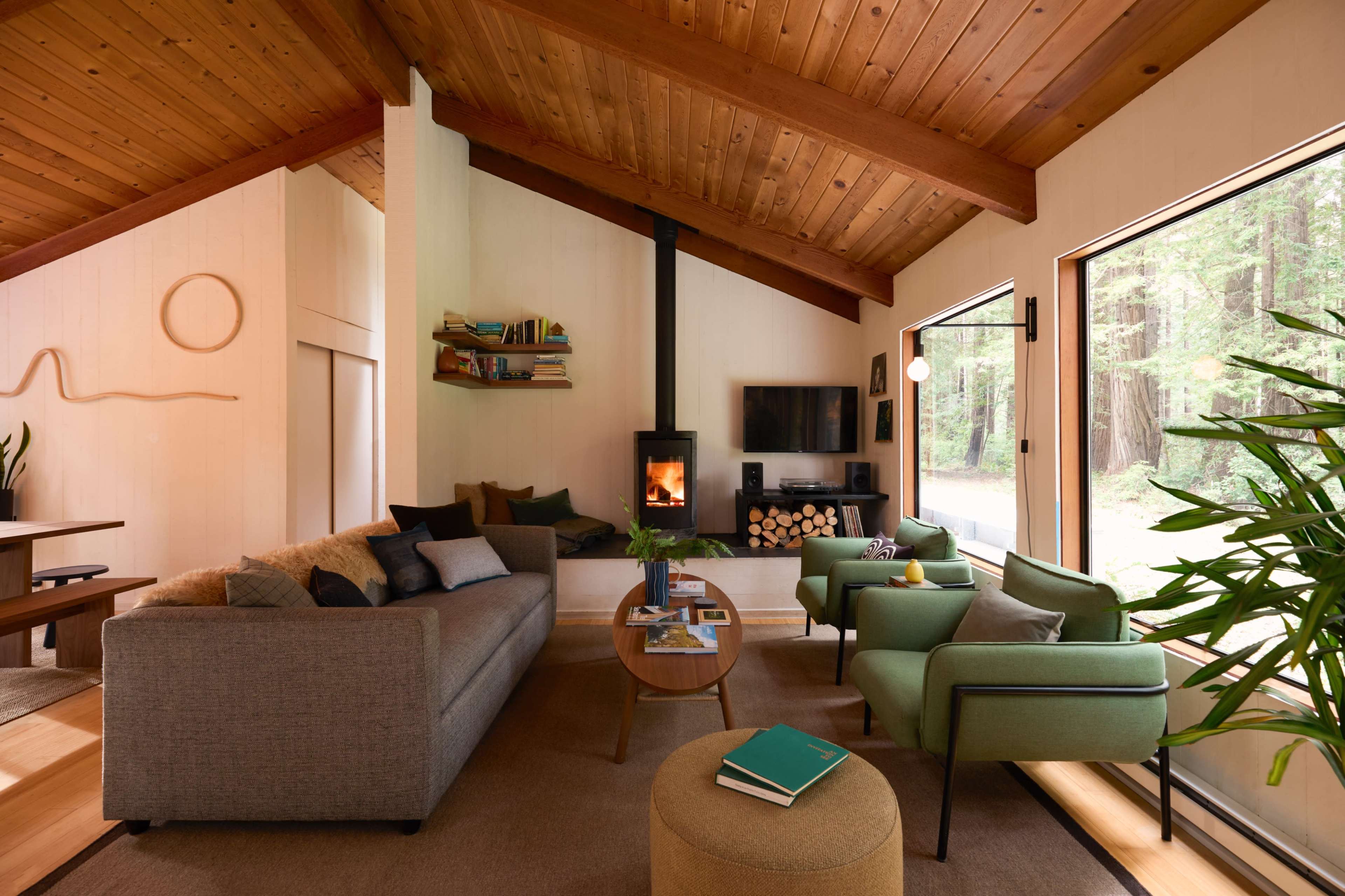The living room features a cozy seating arrangement with a wooden ceiling and large windows overlooking a wooded area, complemented by a modern fireplace and natural decor.