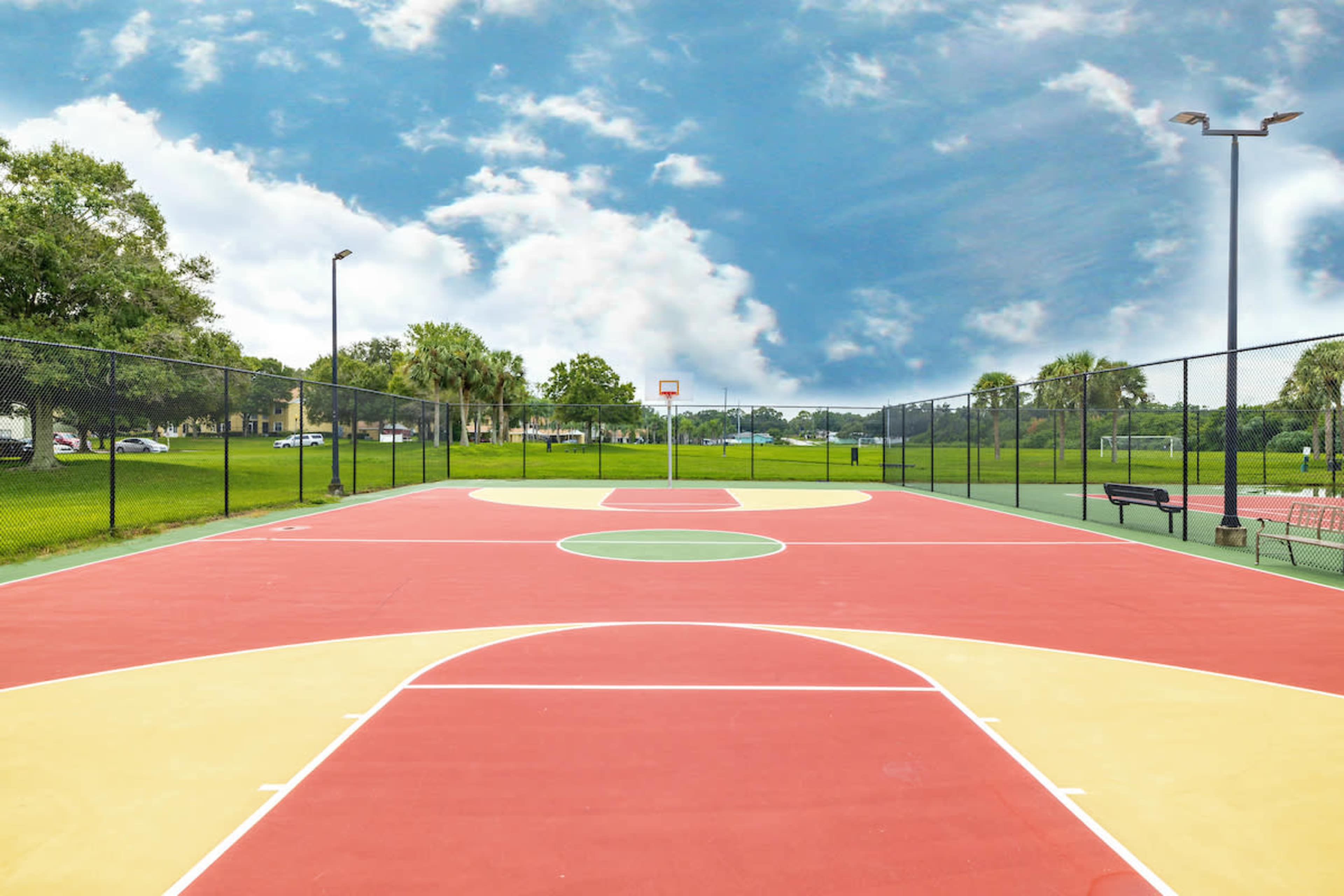 A vacant outdoor basketball court surrounded by grass and trees under a cloudy sky.