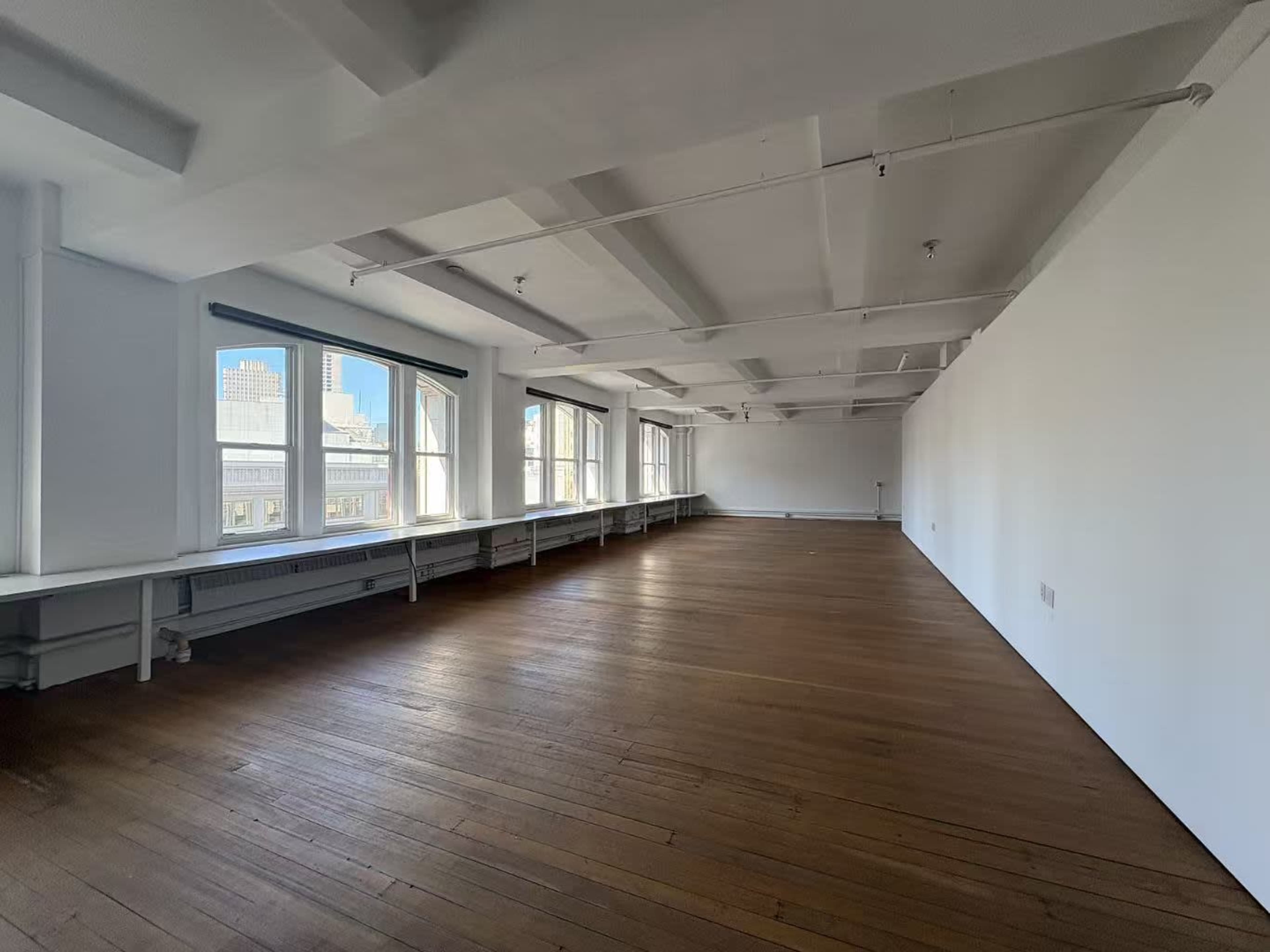 The image shows a spacious, empty room with wooden floors and large windows along one side, letting in natural light.