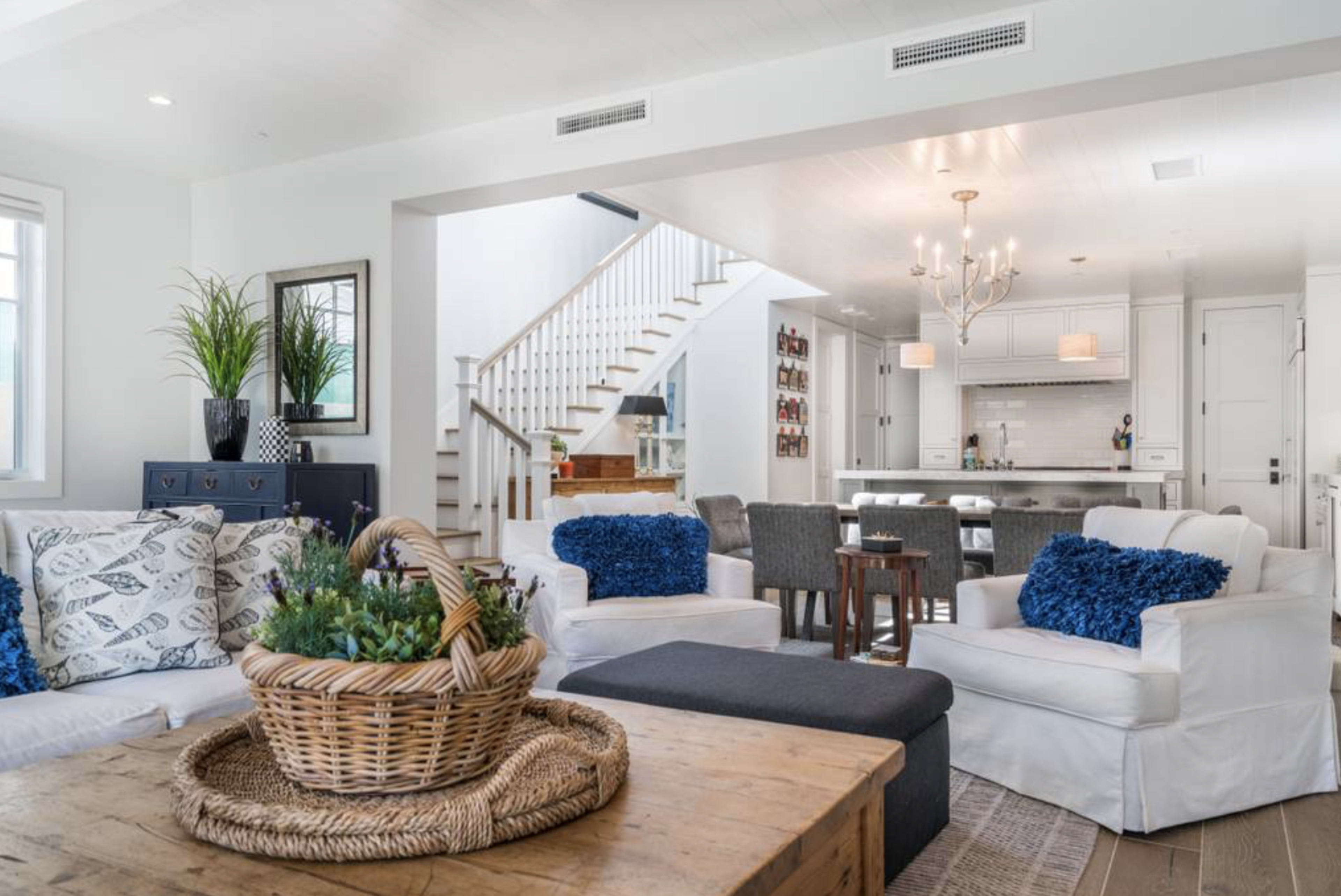 A bright living room features white sofas adorned with blue cushions, a wooden coffee table, and a staircase leading to a kitchen area in the background.