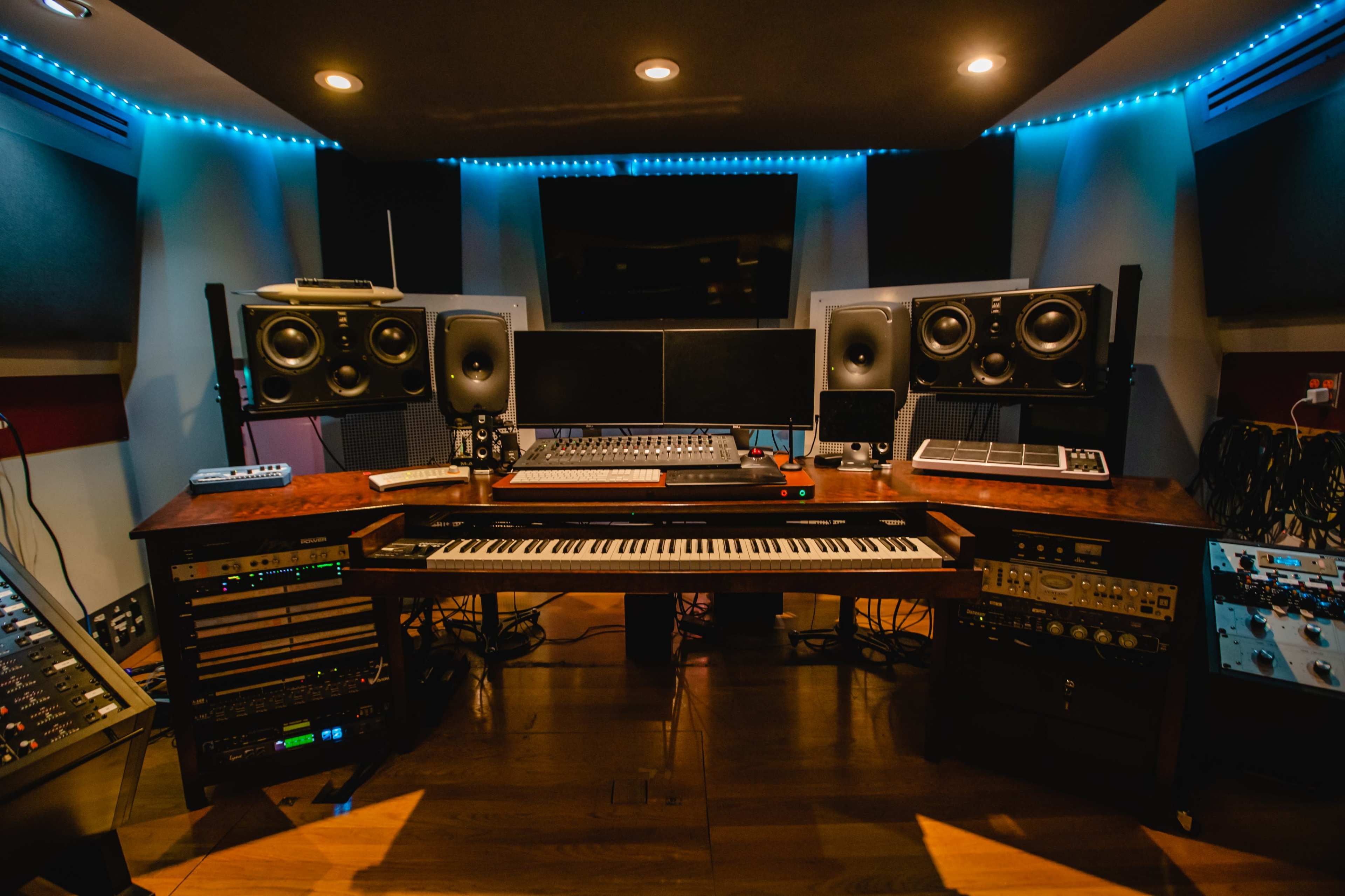 A modern music studio with a central keyboard, two computer monitors, and various audio equipment arranged around a wooden desk.