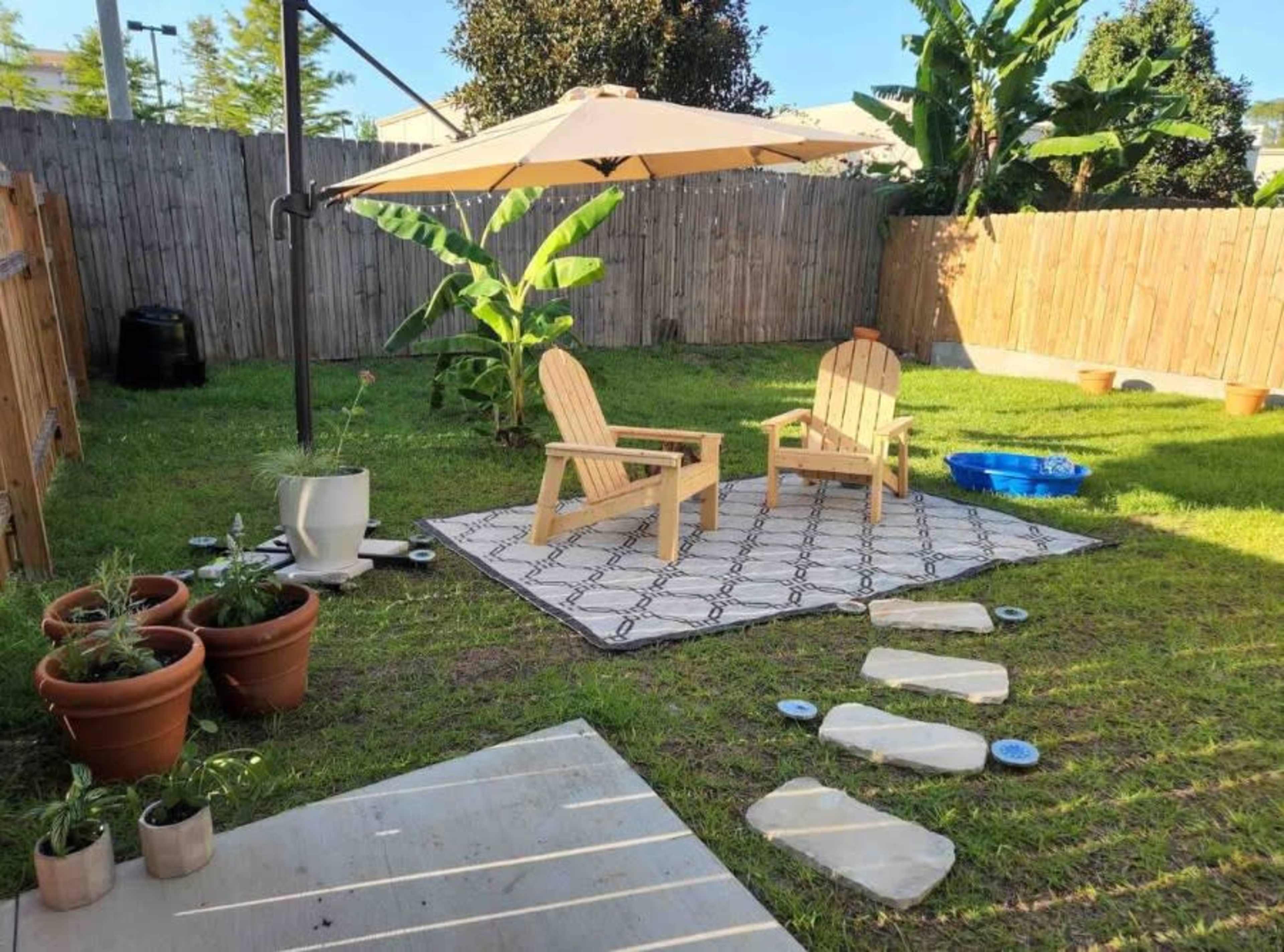 A small backyard features two wooden chairs under an umbrella, surrounded by potted plants, a blue water container, and a grass area with stepping stones.
