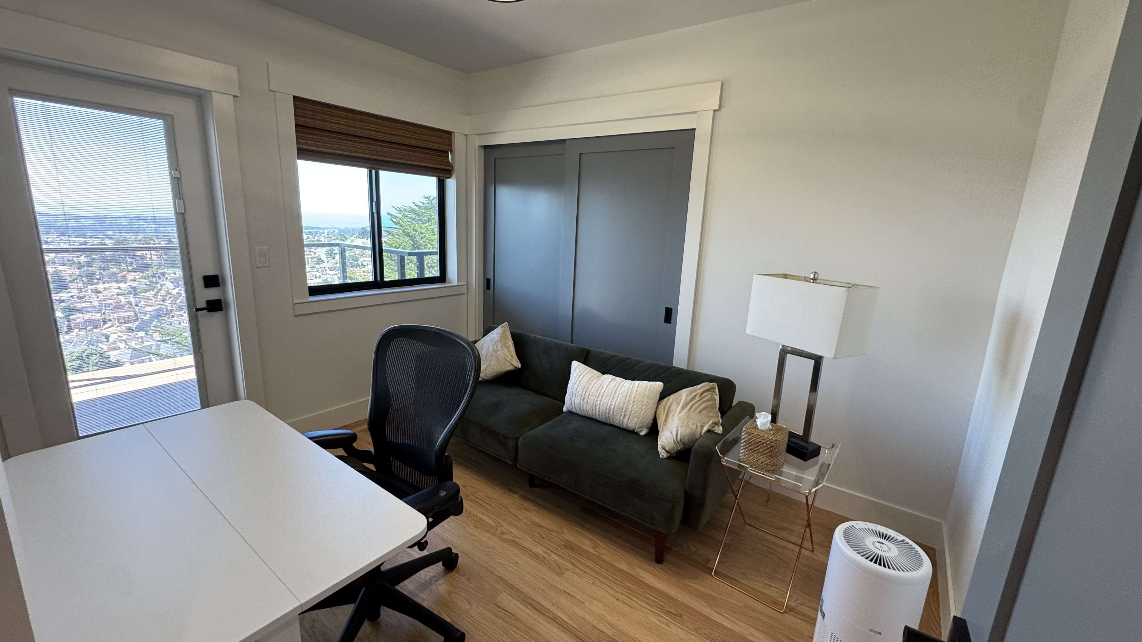 The image shows a small room featuring a desk, a green couch with decorative pillows, a lamp, and a view of the outdoors through a window.