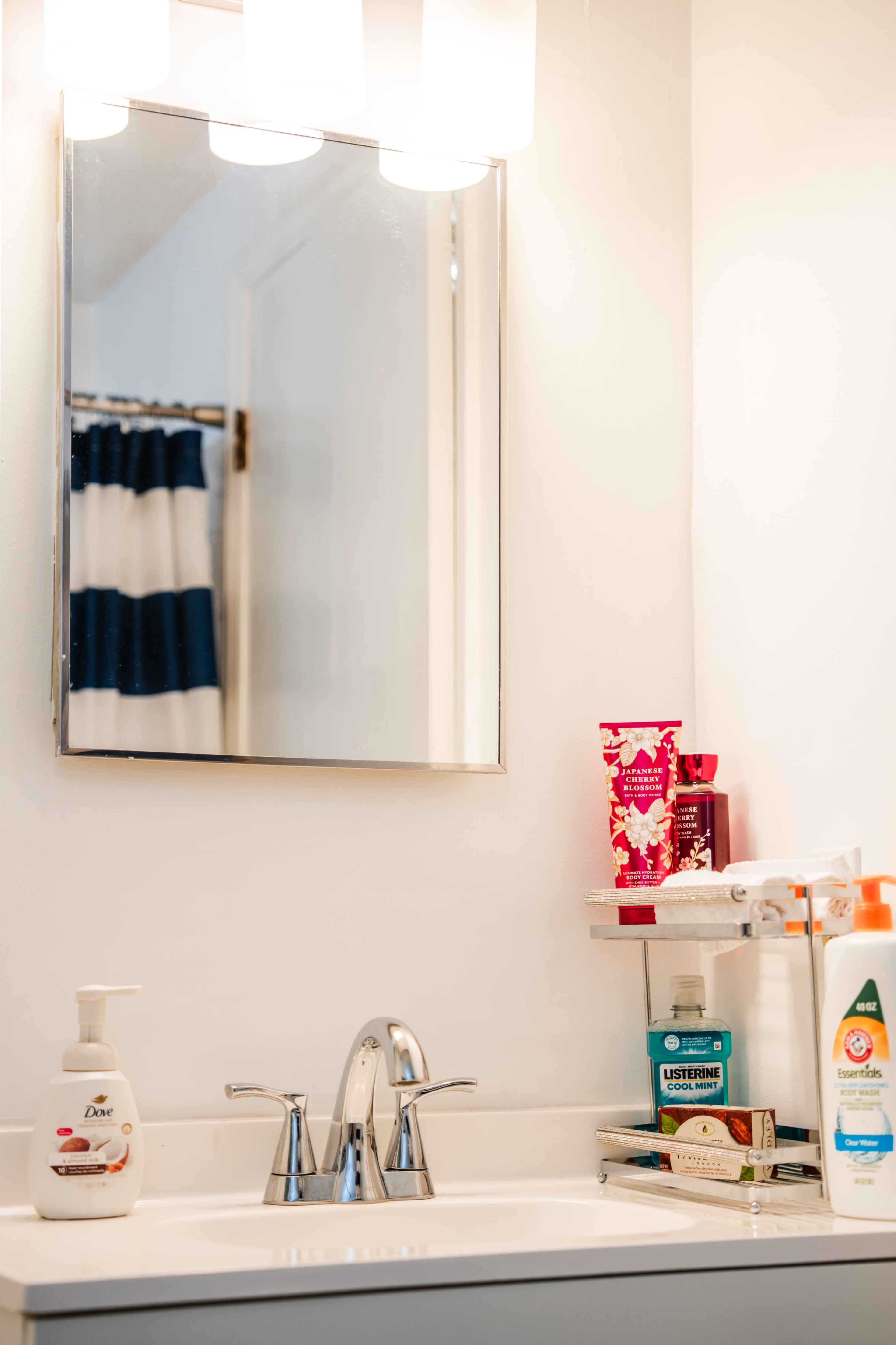 A bathroom sink area features a mirror above the sink, with various toiletry products arranged on the counter and a shower curtain in the background.