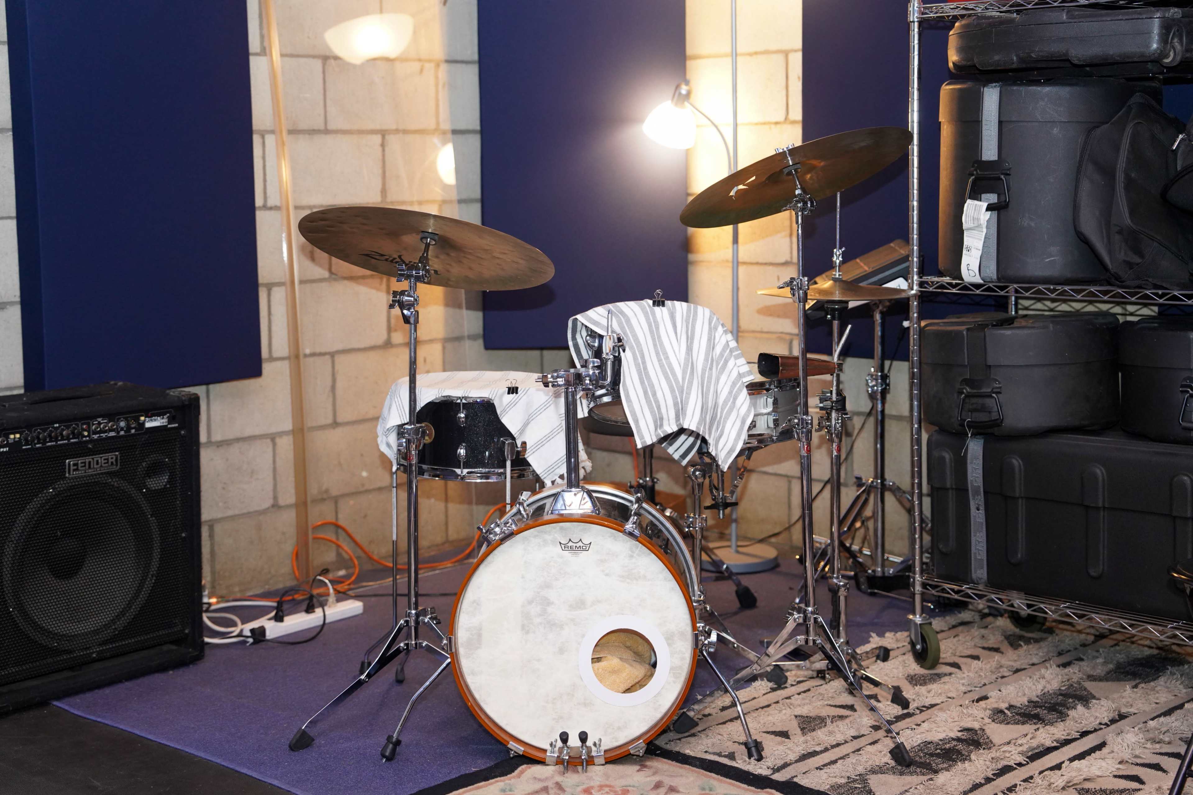 A drum set with cymbals is positioned against a wall in a studio space, alongside amplifiers and storage containers.