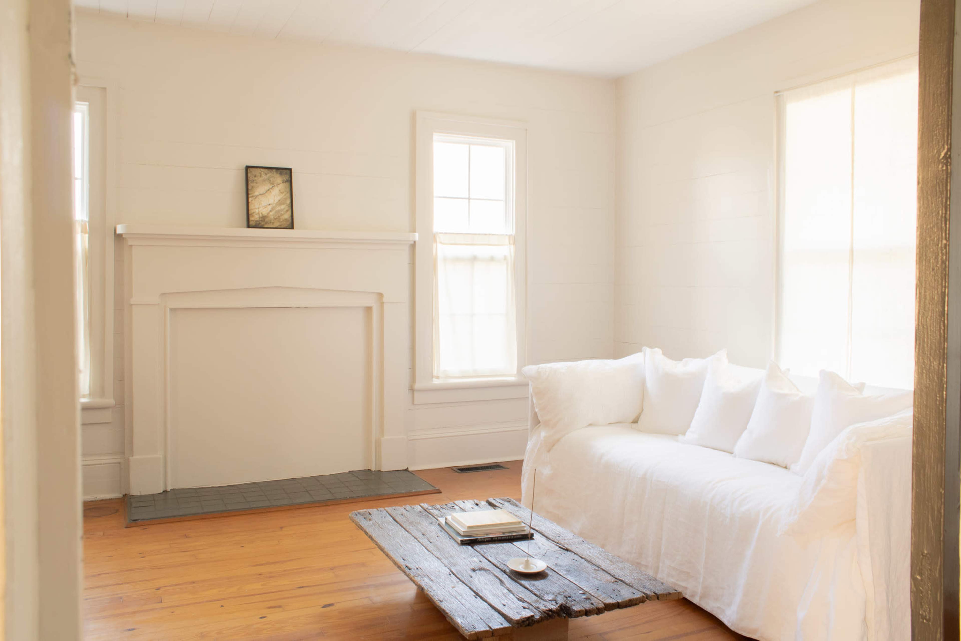 A light-filled room features a white couch with several pillows, a wooden coffee table, and a simple mantel above a fireplace.